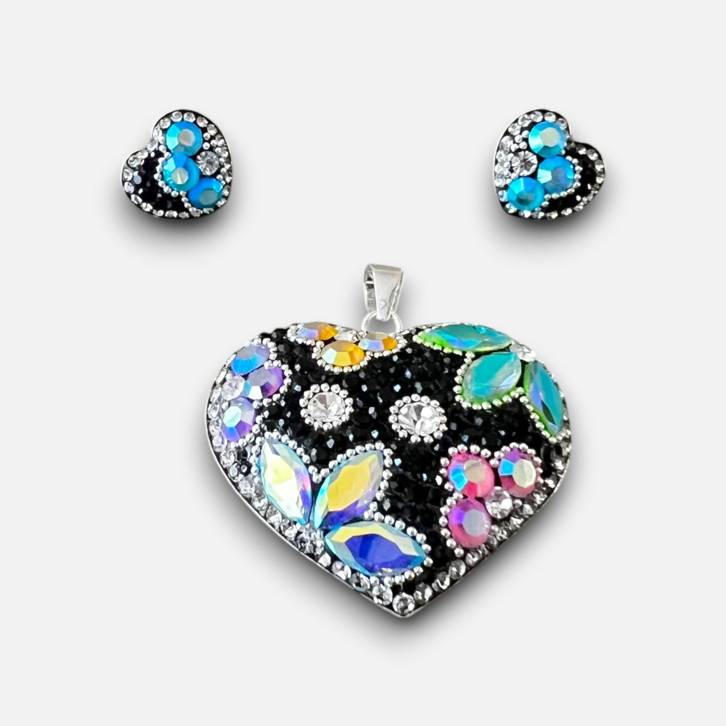 A heart-shaped jewelry set featuring a large pendant and matching stud earrings. The heart design is set in silver with a black background and decorated with iridescent, rainbow-colored rhinestones, blue stones, and clear crystals.