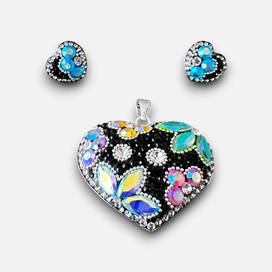A heart-shaped jewelry set featuring a large pendant and matching stud earrings. The heart design is set in silver with a black background and decorated with iridescent, rainbow-colored rhinestones, blue stones, and clear crystals.