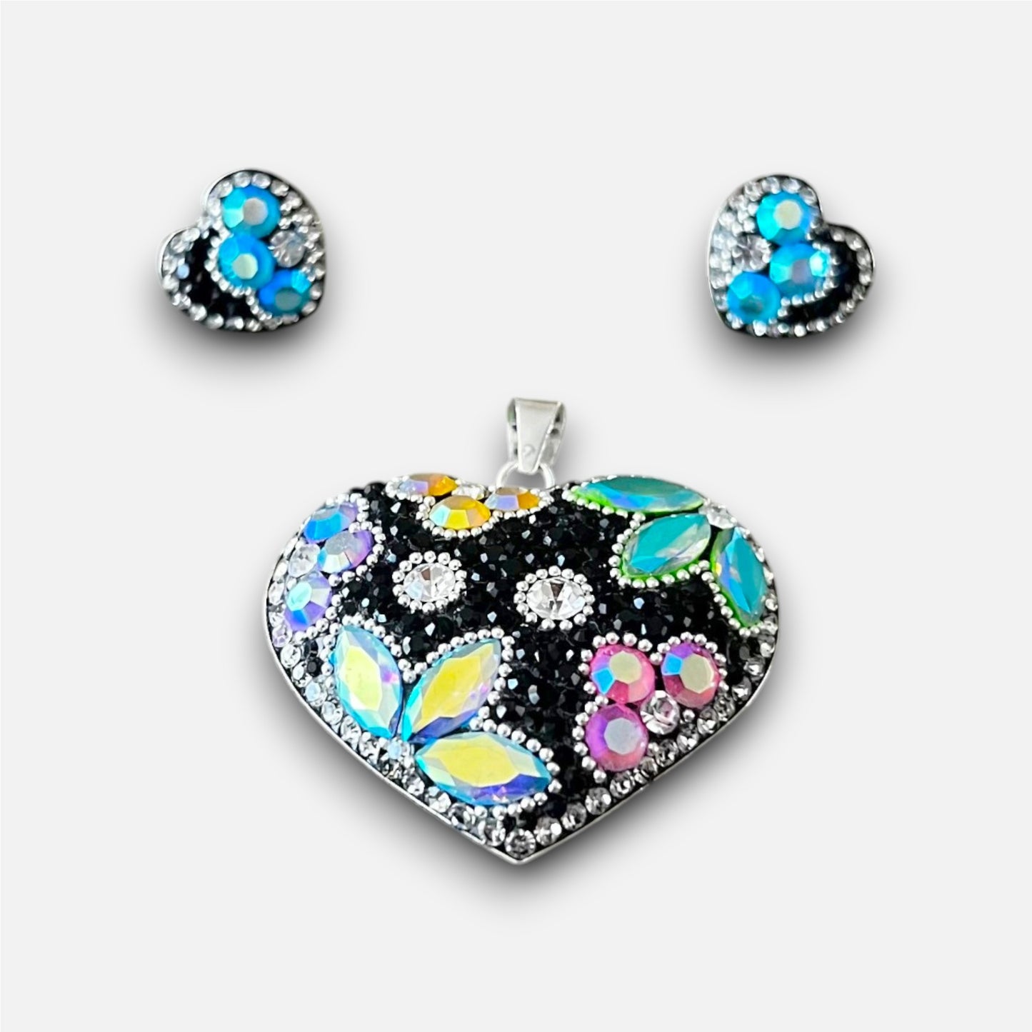A heart-shaped jewelry set featuring a large pendant and matching stud earrings. The pendant and earrings are set in silver-tone metal and decorated with a black textured background, iridescent butterfly wings in blue, yellow, and green, and small colorful floral clusters.