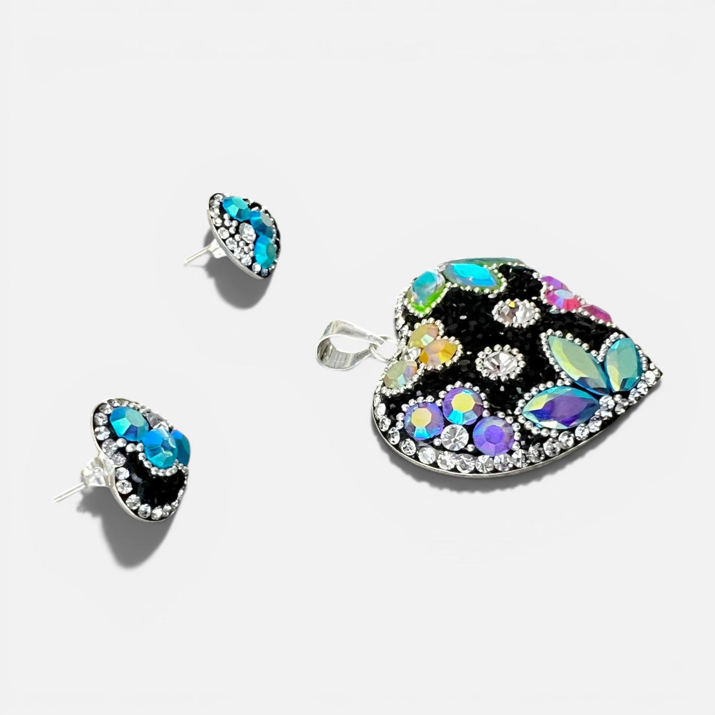 A jewelry set featuring a large heart-shaped pendant and two matching stud earrings. The heart pendant has a black background decorated with iridescent, multicolored rhinestones in various shapes, surrounded by small clear rhinestones on a silver-tone setting.