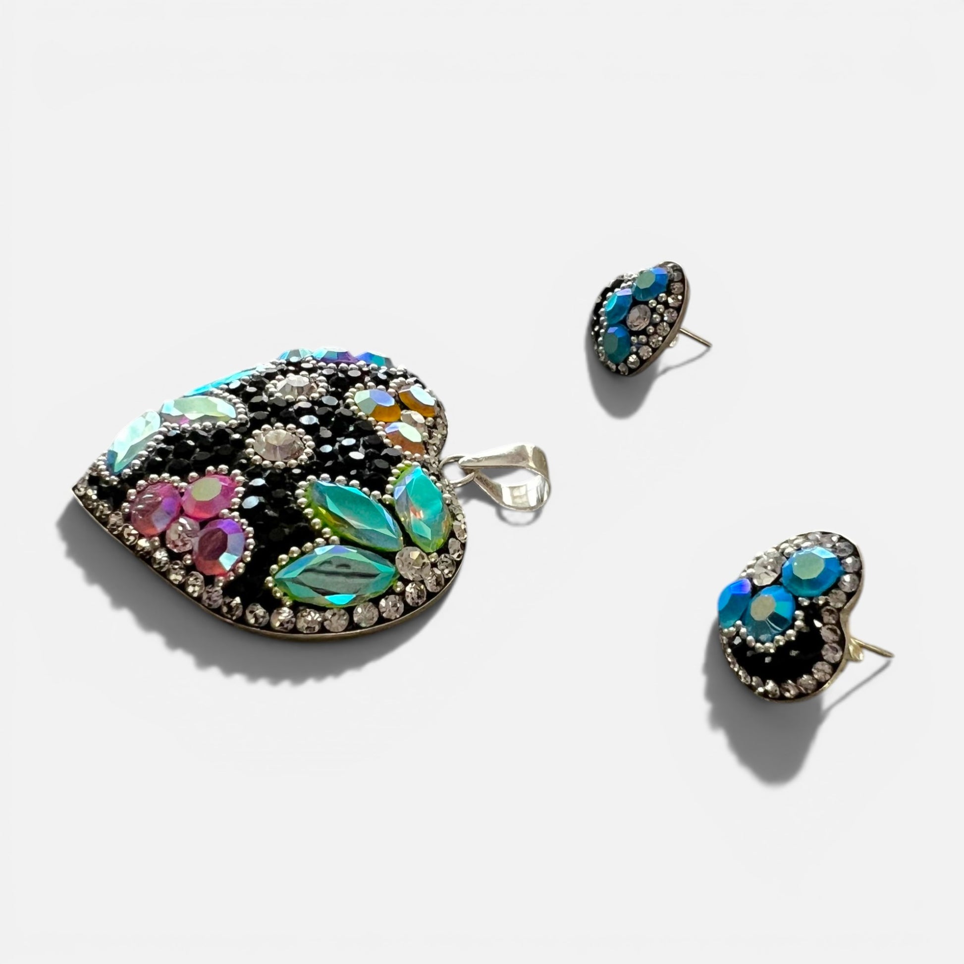 A heart-shaped statement brooch and matching stud earrings featuring a black background embellished with iridescent turquoise, green, pink, and blue stones, accented by clear rhinestones on a light gray background.
