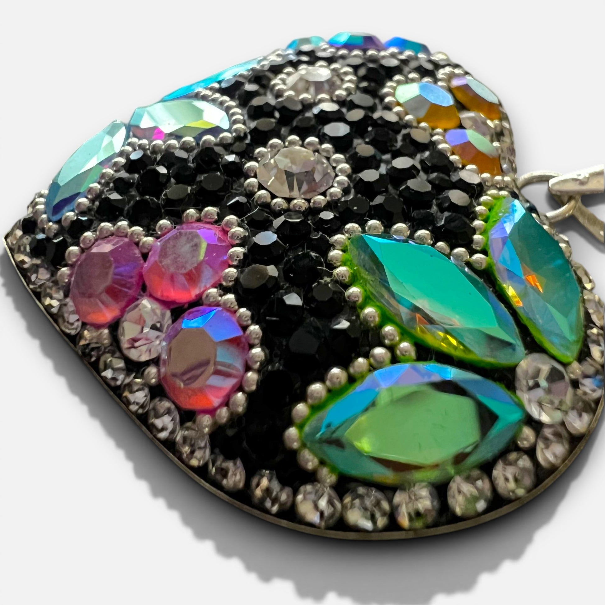 A close-up of a heart-shaped embellished clutch purse featuring a black rhinestone base decorated with iridescent green, pink, and blue crystals, silver rhinestones, and a gold-tone metal frame.