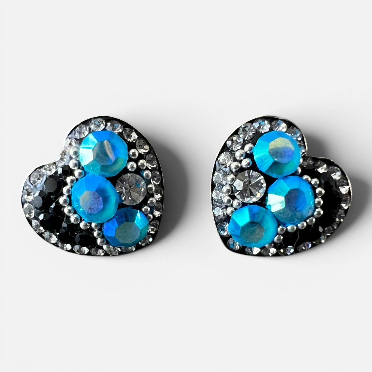 A pair of heart-shaped stud earrings featuring vibrant blue and teal iridescent rhinestones, accented by black and silver crystals on a light gray background.