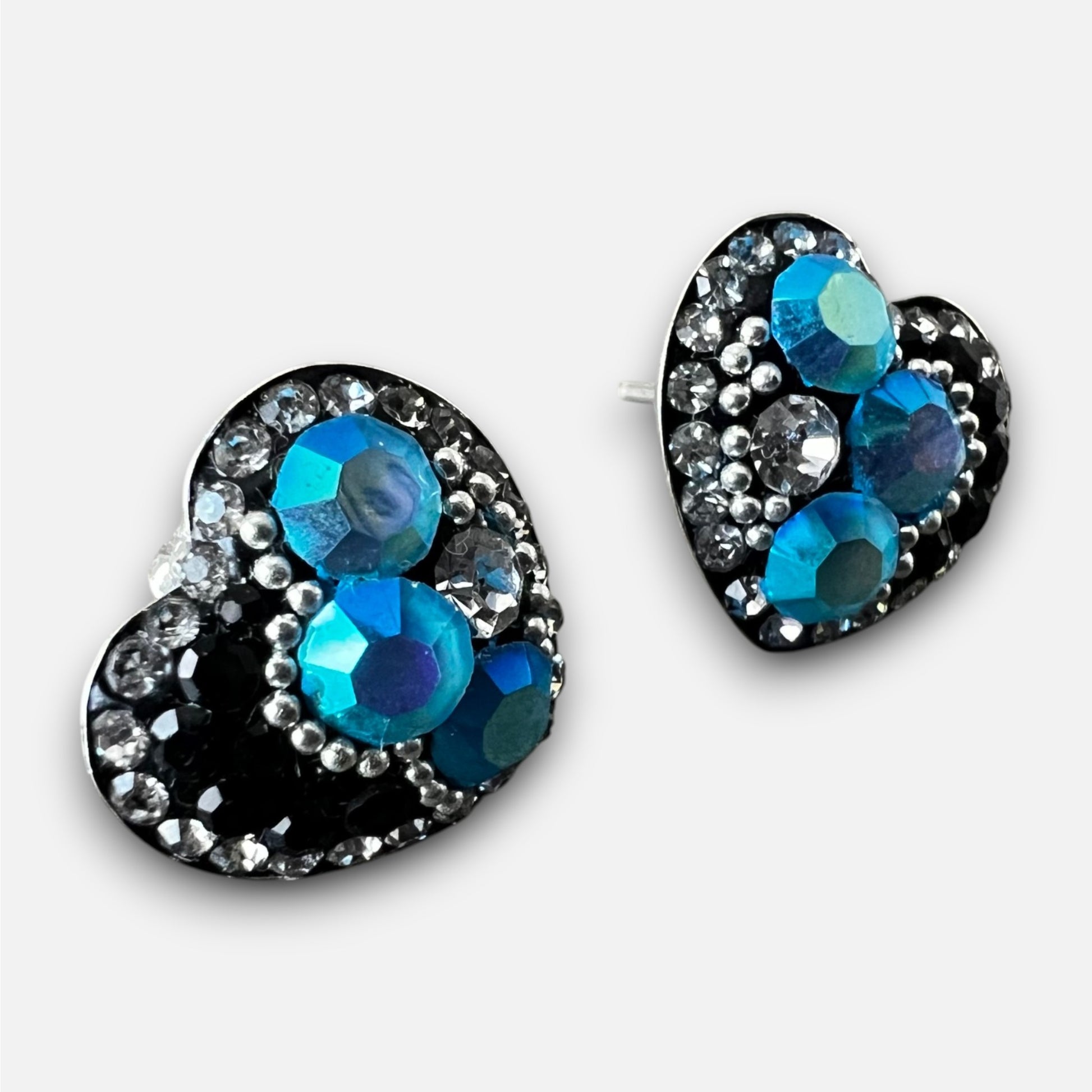 A pair of heart-shaped stud earrings featuring large faceted blue and teal rhinestones surrounded by smaller silver and clear rhinestones on a black background, shown on a light gray studio background.