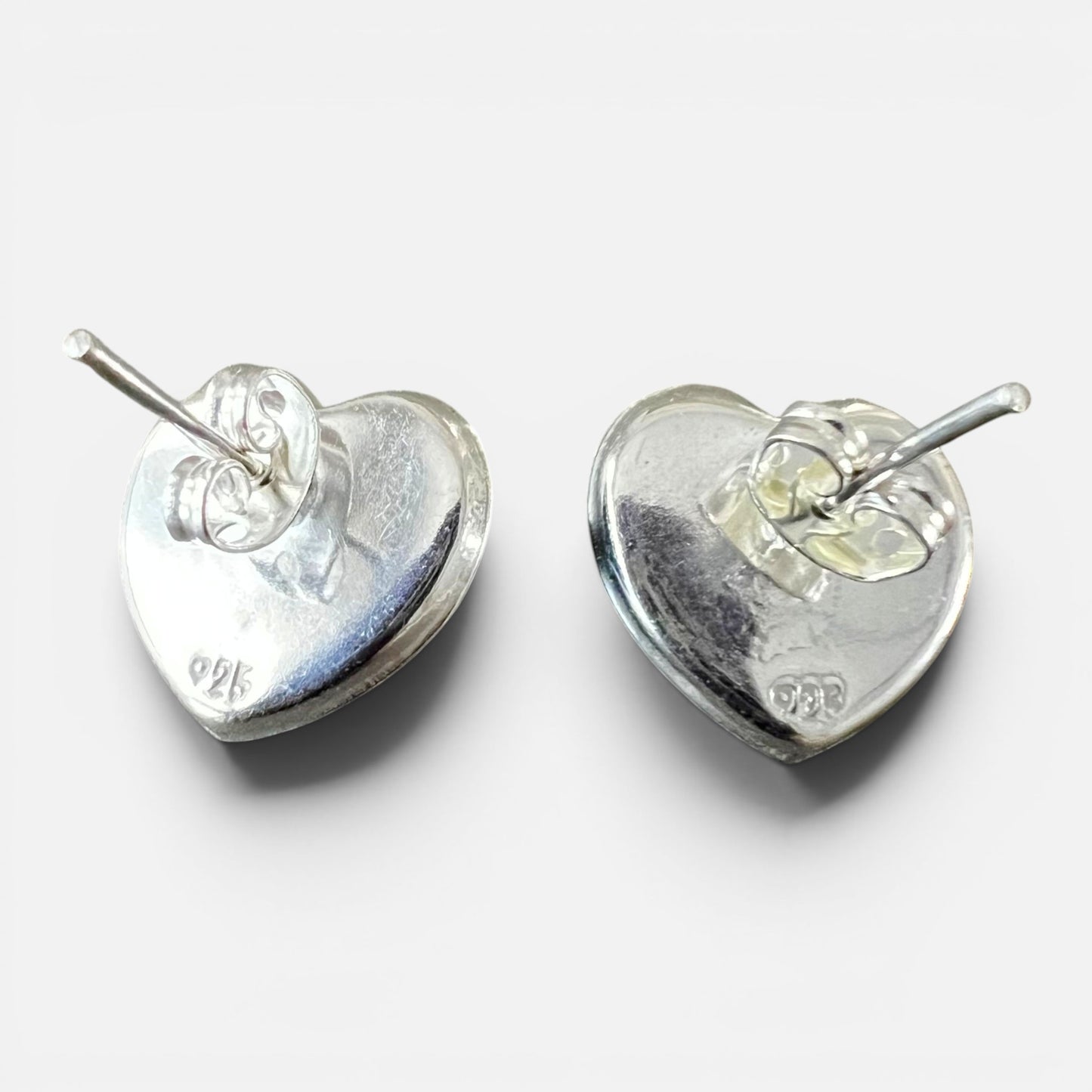A pair of sterling silver heart-shaped stud earrings shown from the back, displaying the polished metal surfaces and post settings against a light gray background. Each earring has a 925 stamp.