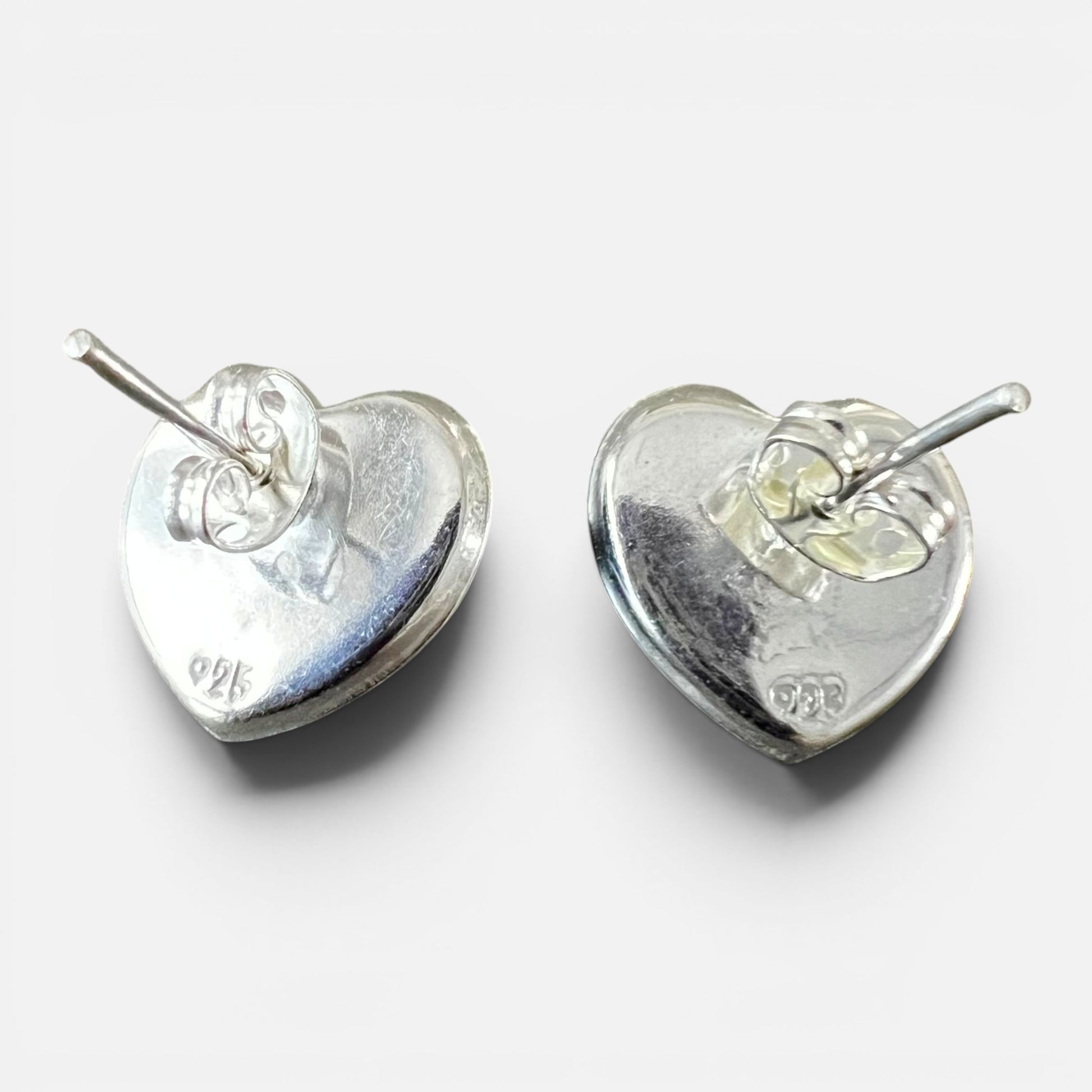 A pair of sterling silver heart-shaped stud earrings shown from the back, displaying the polished metal surfaces and post settings against a light gray background. Each earring has a 925 stamp.