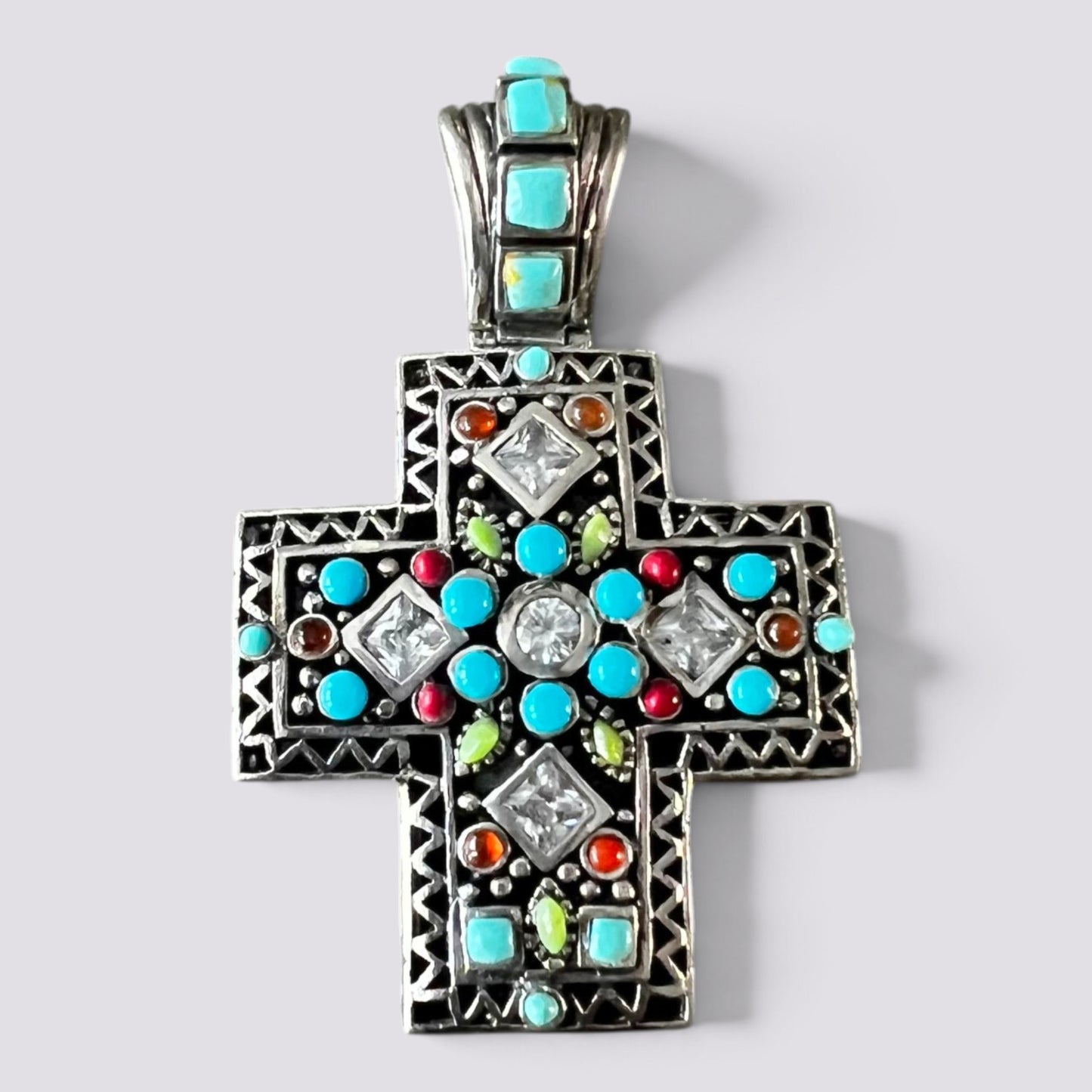 A silver-toned cross pendant featuring turquoise, red, green, and orange stones, with clear rhinestones, set against a light gray background.