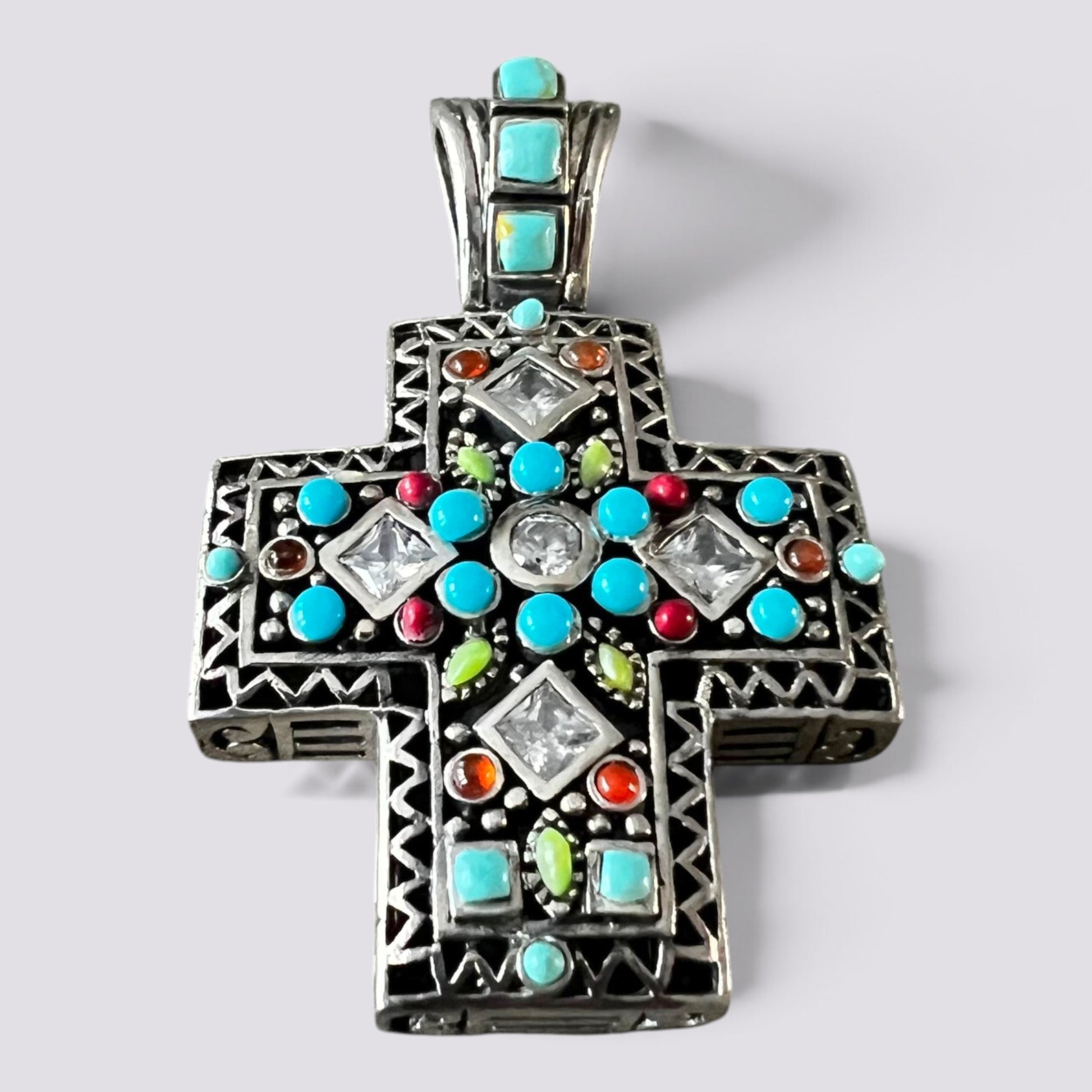 An ornate sterling silver cross pendant featuring multiple inlaid turquoise, red, green, and clear gemstones, photographed on a neutral light gray background.