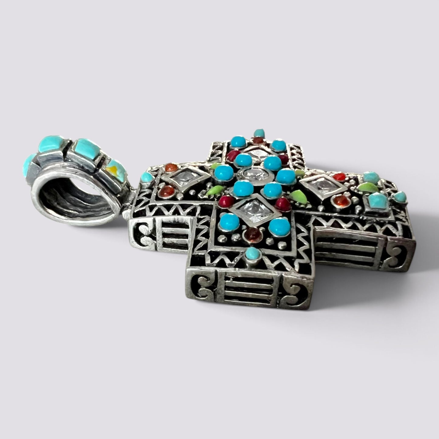 An ornate silver cross pendant decorated with turquoise, red, and green stones, shown on a light gray background.