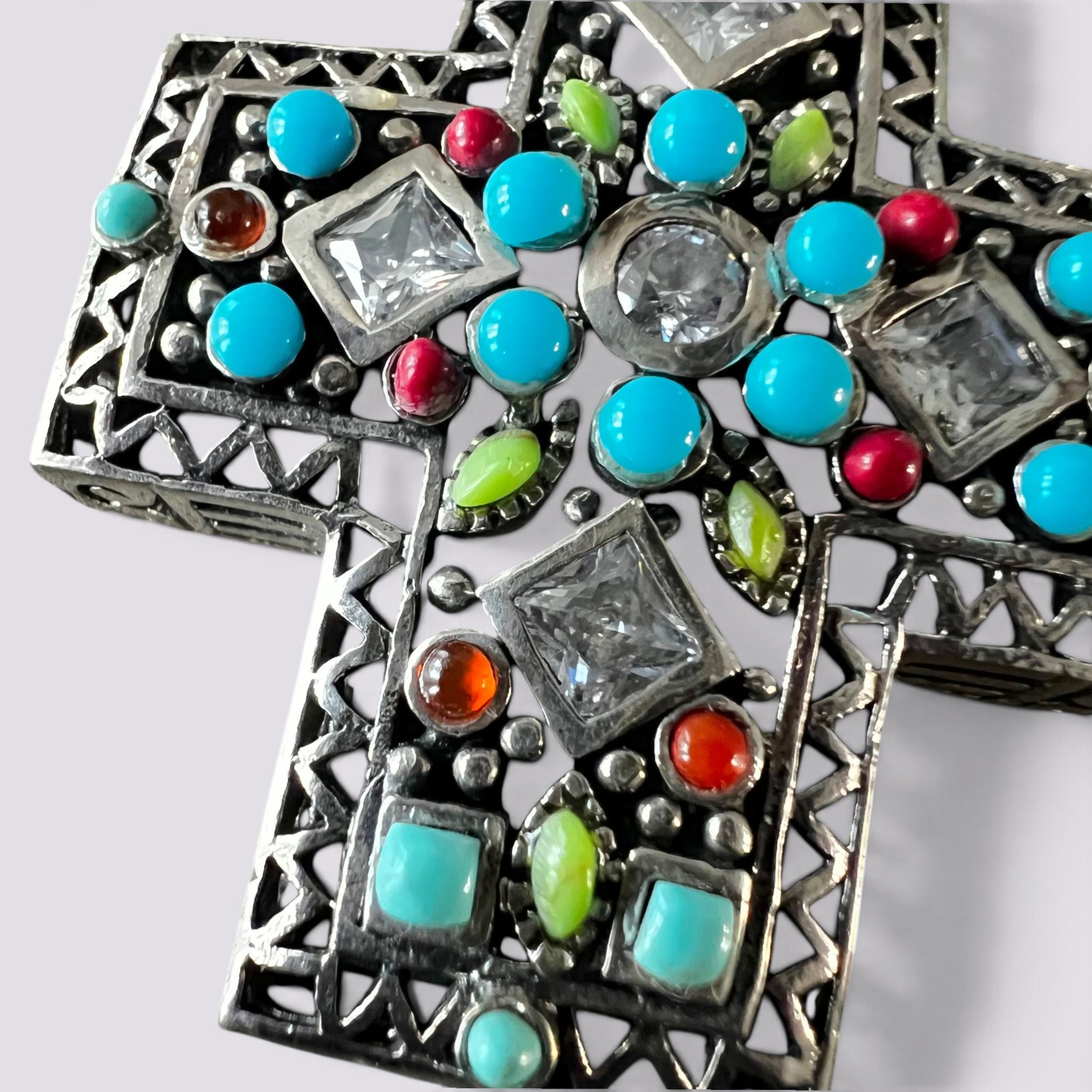 An ornate silver filigree cross pendant decorated with turquoise, red, green, and orange stones, shown on a light gray background.