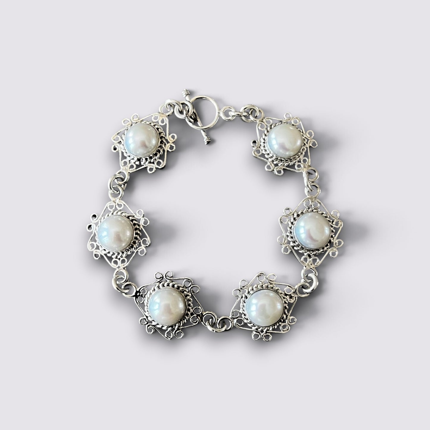 A silver-toned bracelet featuring five ornate filigree settings with round white faux pearls, arranged in a circle on a light gray background.
