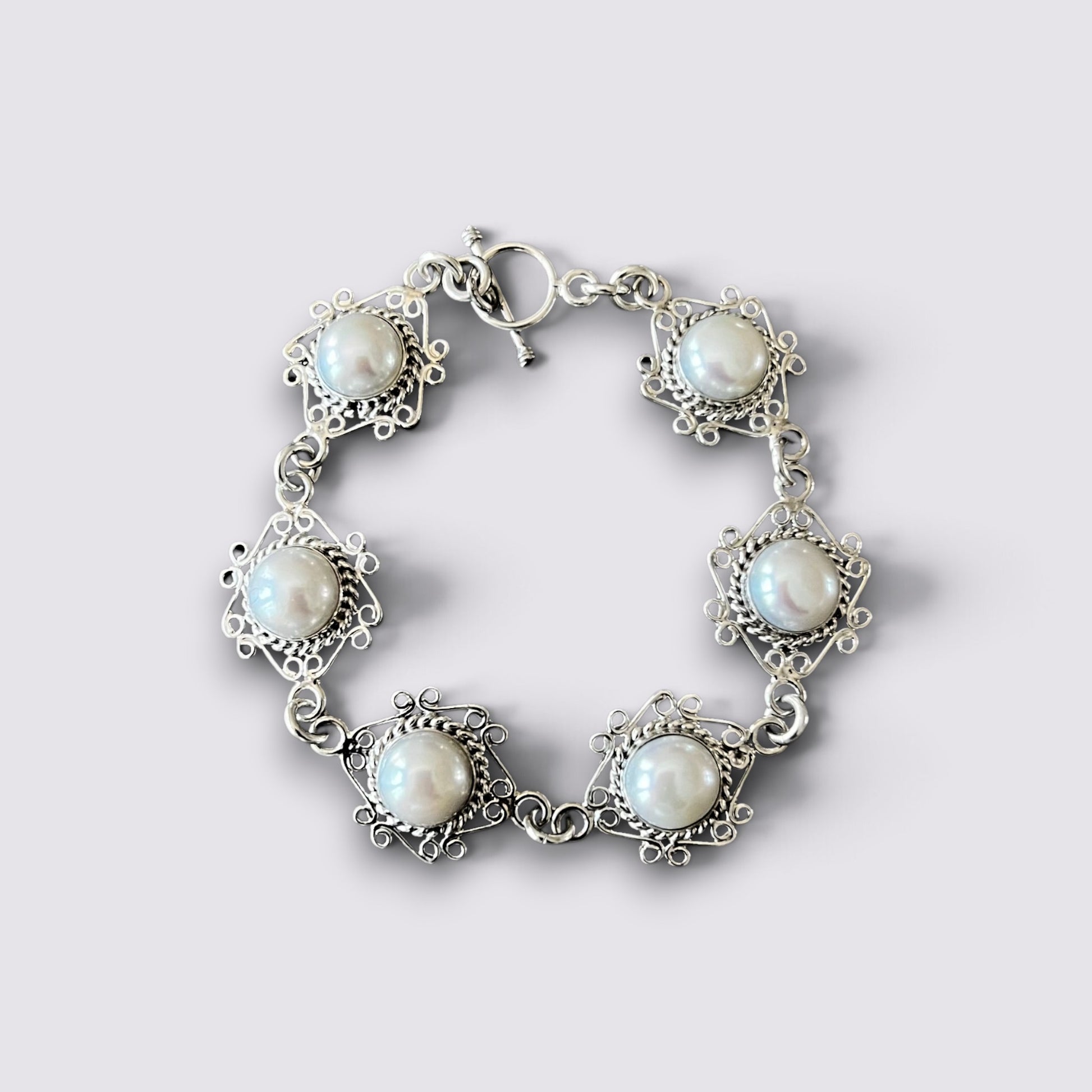 A silver-toned bracelet featuring five ornate filigree settings with round white faux pearls, arranged in a circle on a light gray background.