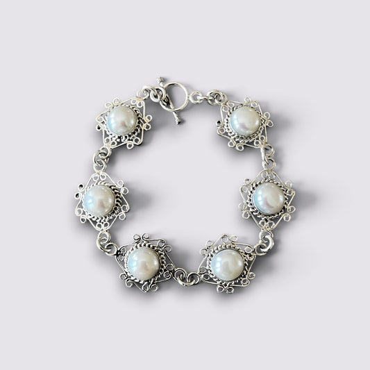 A silver-toned bracelet featuring five ornate filigree settings with round white faux pearls, arranged in a circle on a light gray background.
