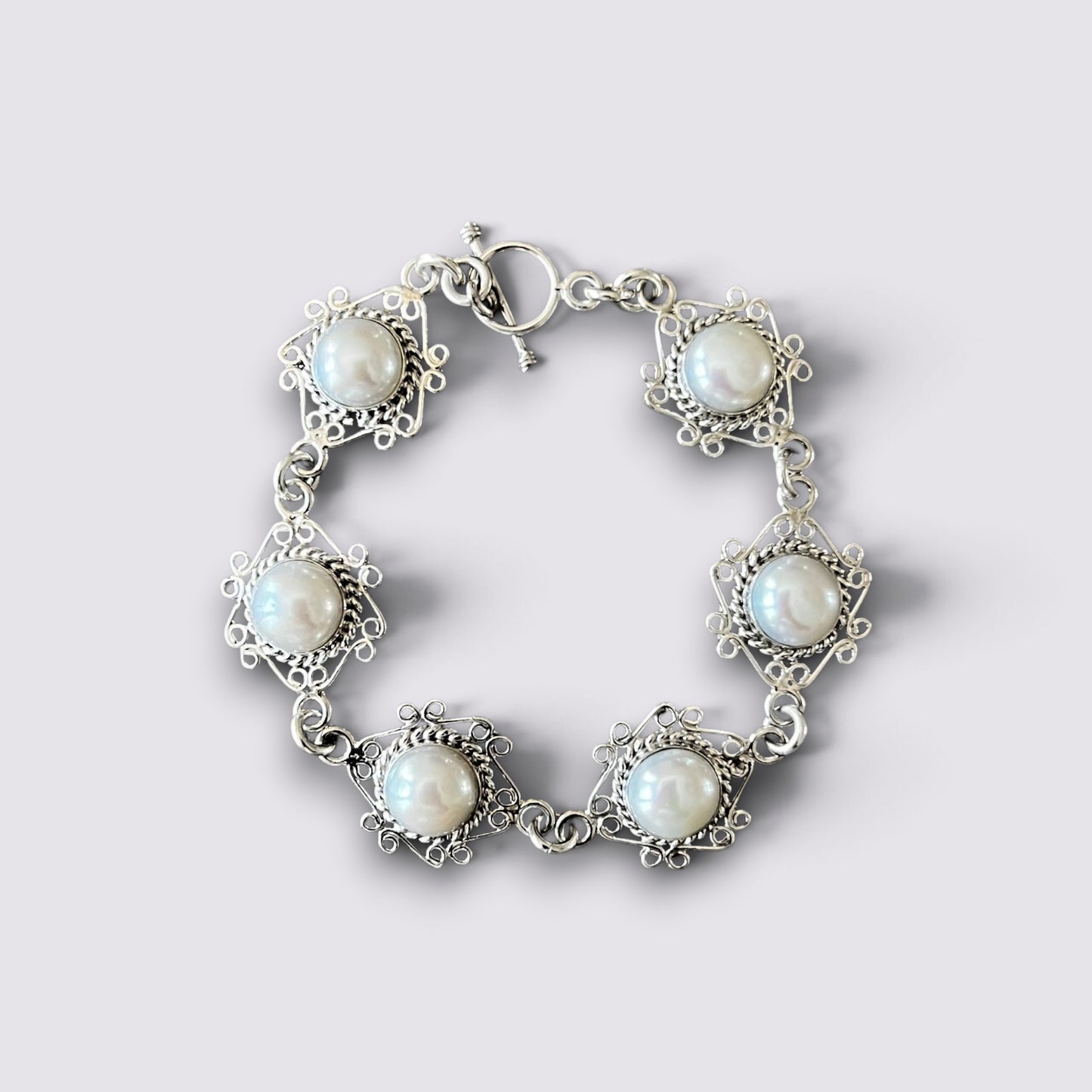 A silver-tone bracelet featuring five ornate floral settings, each holding a round white pearl, arranged in a circle on a light gray background.