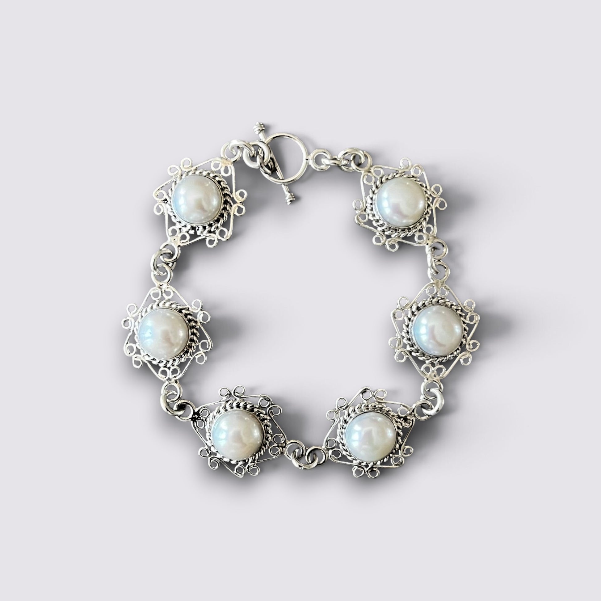 A silver-tone bracelet featuring five ornate floral settings, each holding a round white pearl, arranged in a circle on a light gray background.
