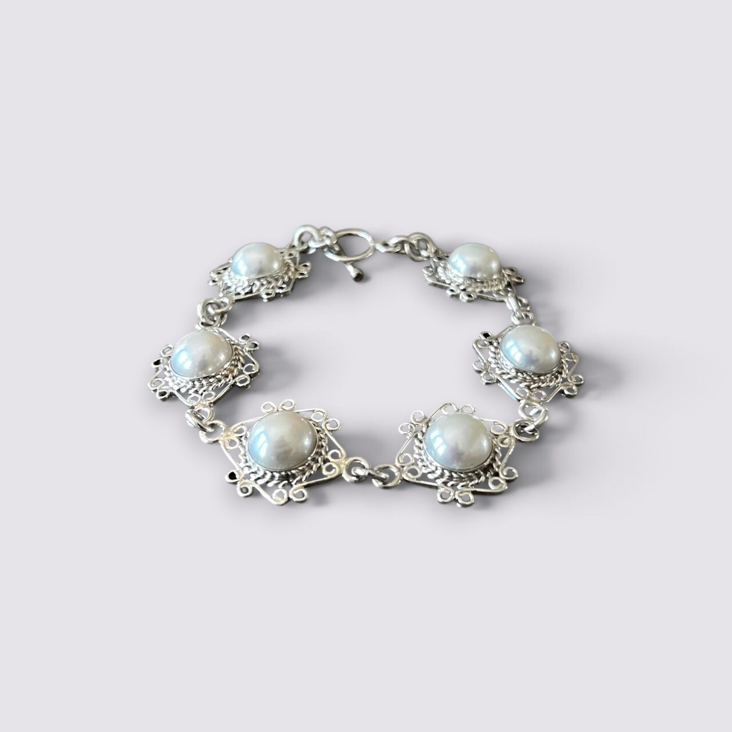 A silver-tone bracelet featuring five round white faux pearls set in ornate filigree frames, shown on a light gray background.