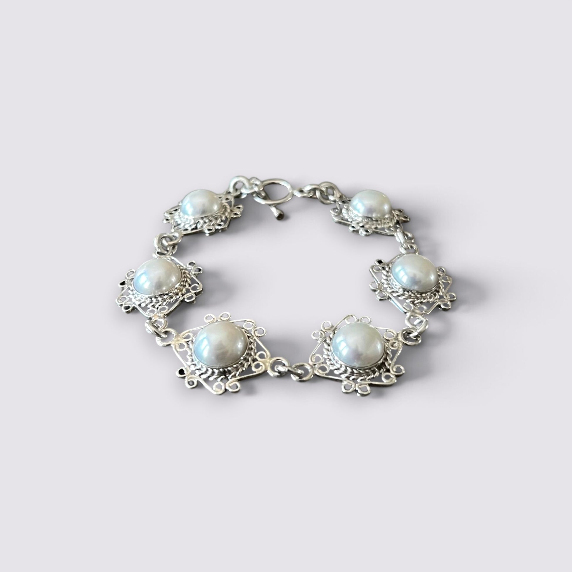 A silver-tone bracelet featuring five round white faux pearls set in ornate filigree frames, shown on a light gray background.