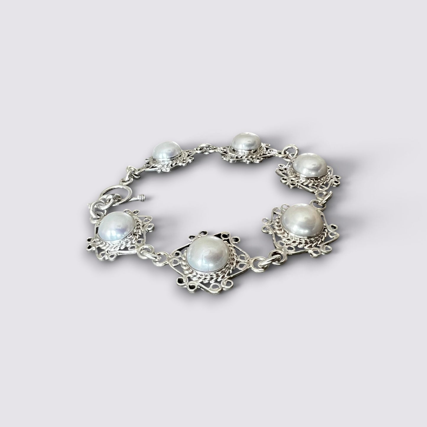 A silver-toned filigree bracelet featuring five round white pearls set in ornate openwork metal frames, shown on a light gray background.