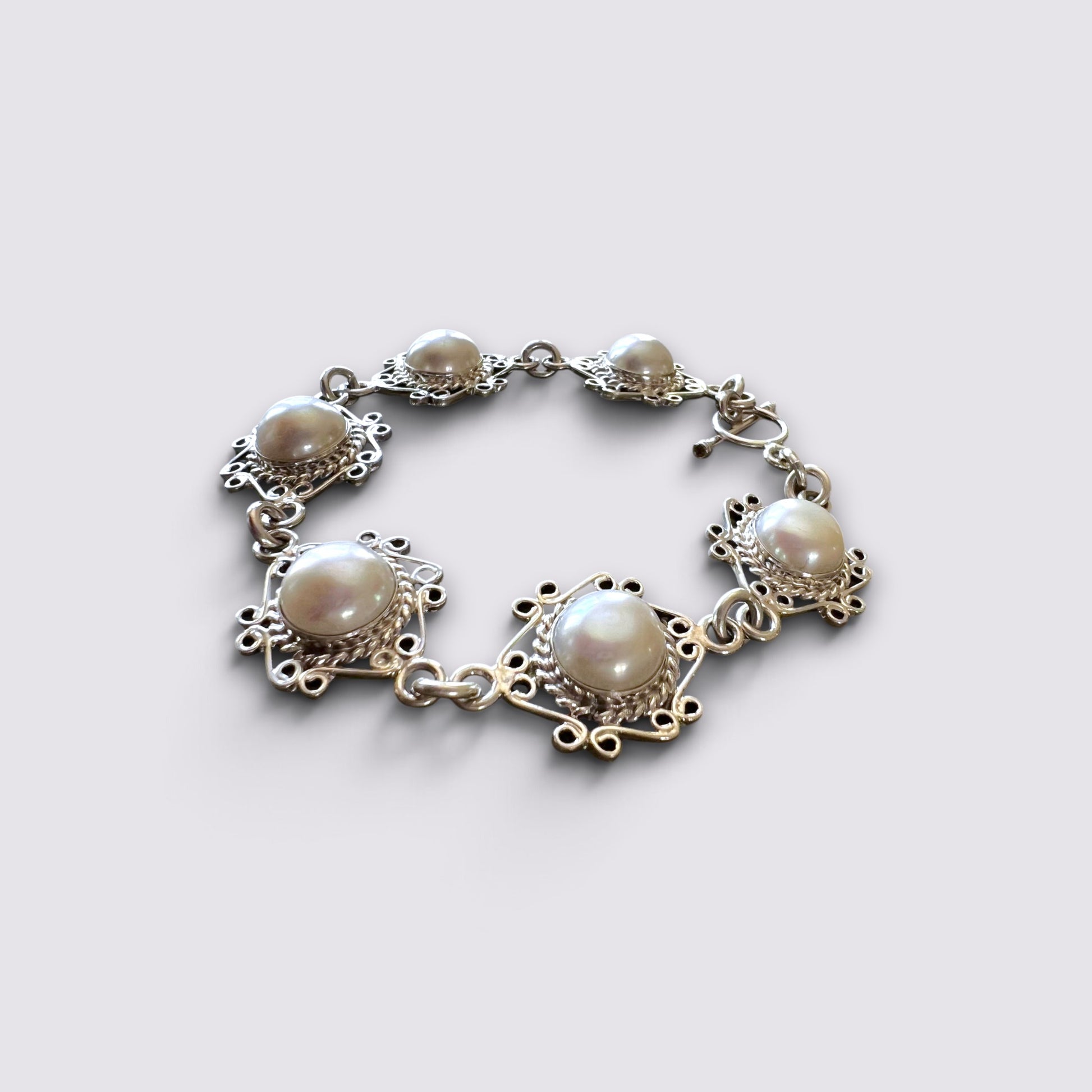 A silver-toned bracelet featuring five large oval pearl-like stones set in ornate filigree settings, arranged in a loose circle on a light gray background.