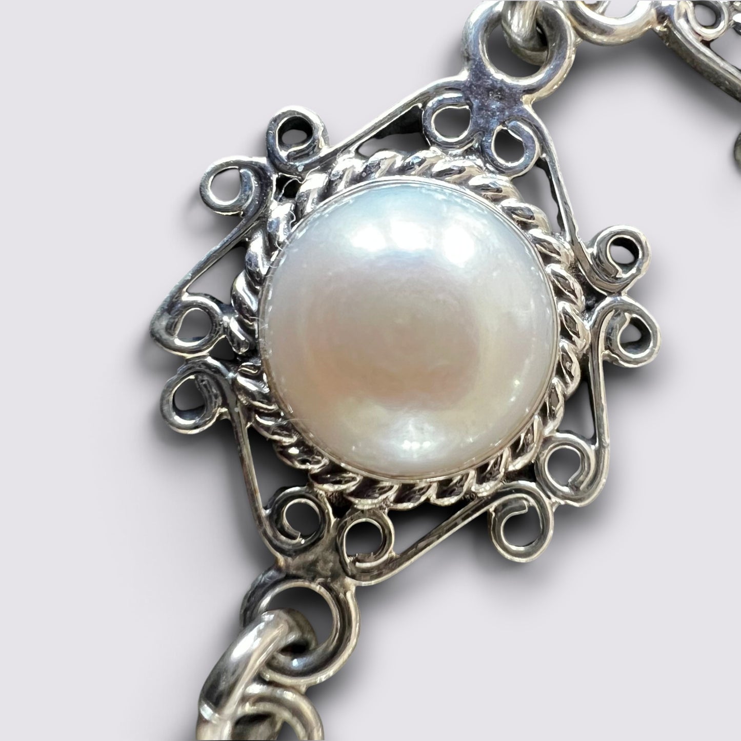 A close-up of a silver-toned filigree jewelry clasp featuring a round, lustrous white pearl-like center on a light gray background.