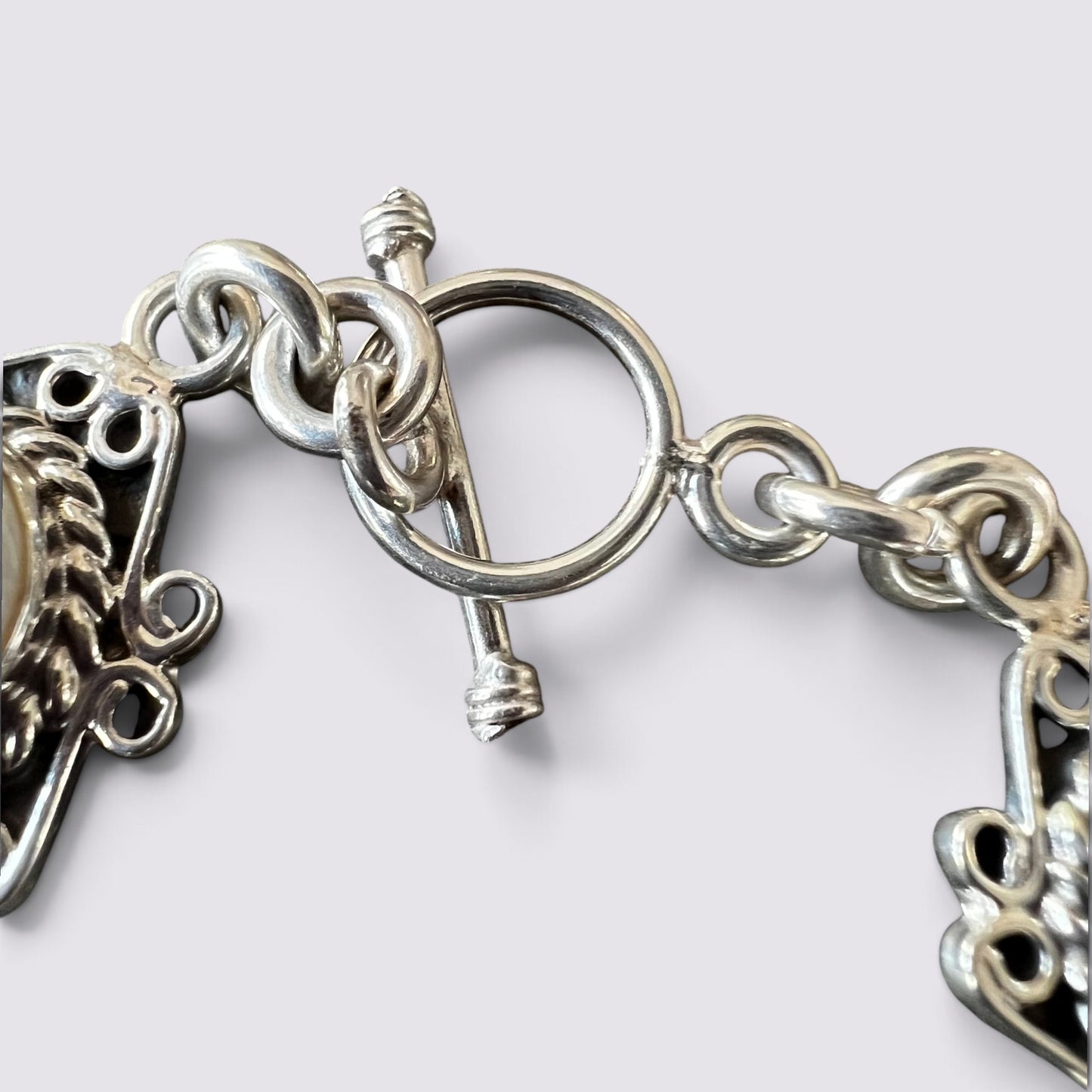 Close-up of a silver-toned chain bracelet with a toggle clasp and ornate decorative links on a light gray background.