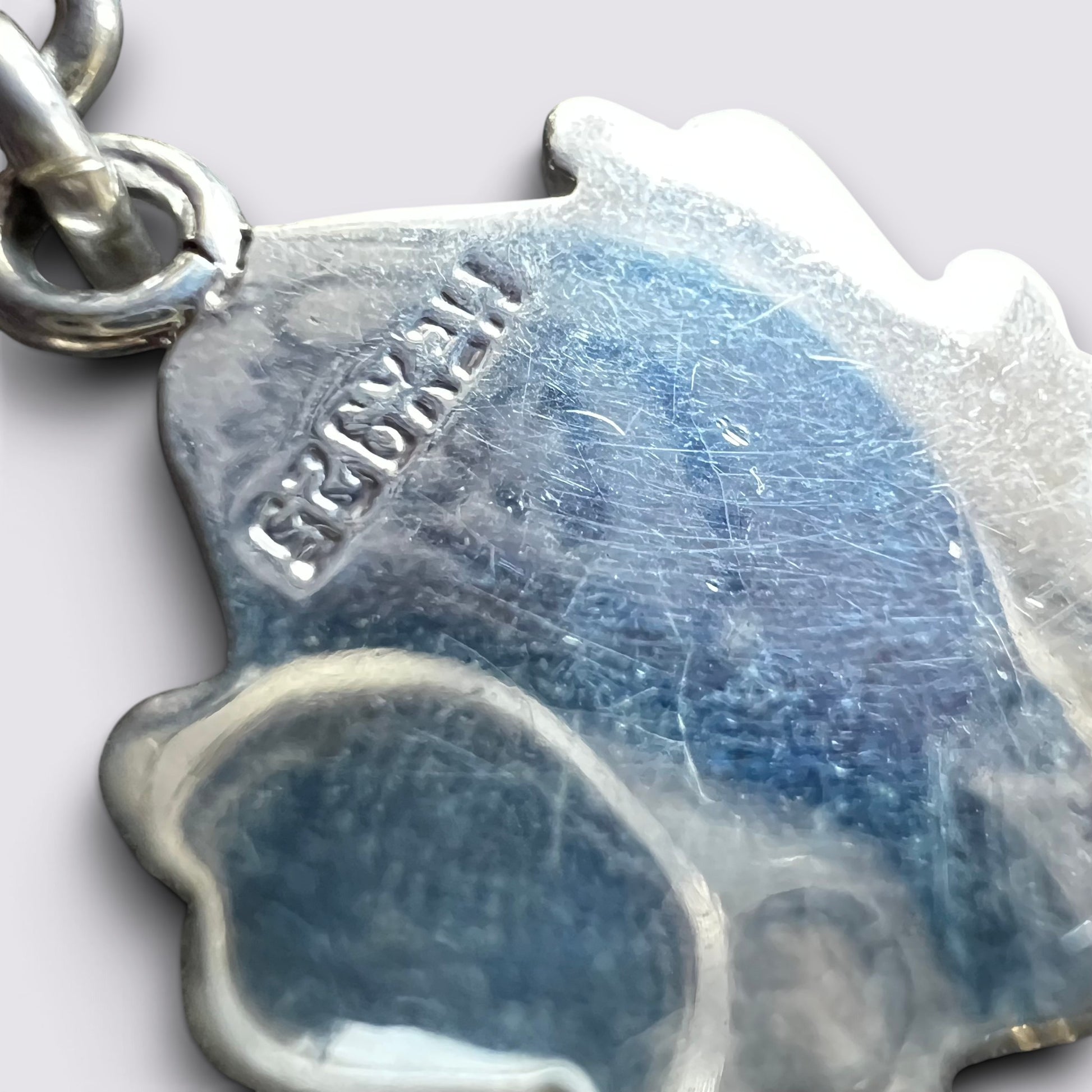 Close-up of a silver dog-shaped pendant charm with a blue inlay, showing the brand name 'HESKULL' stamped on the side and a silver clasp at the top left.