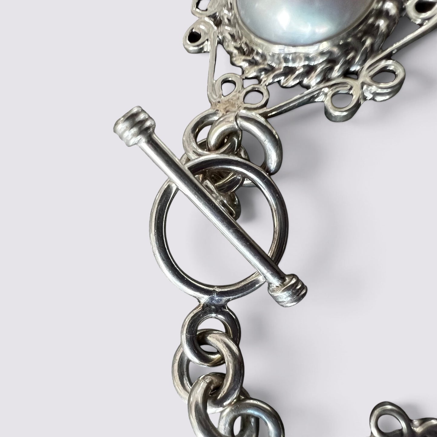 Close-up of a silver-toned chain bracelet featuring a toggle clasp and a round iridescent pearl at the top, shown against a light gray background.