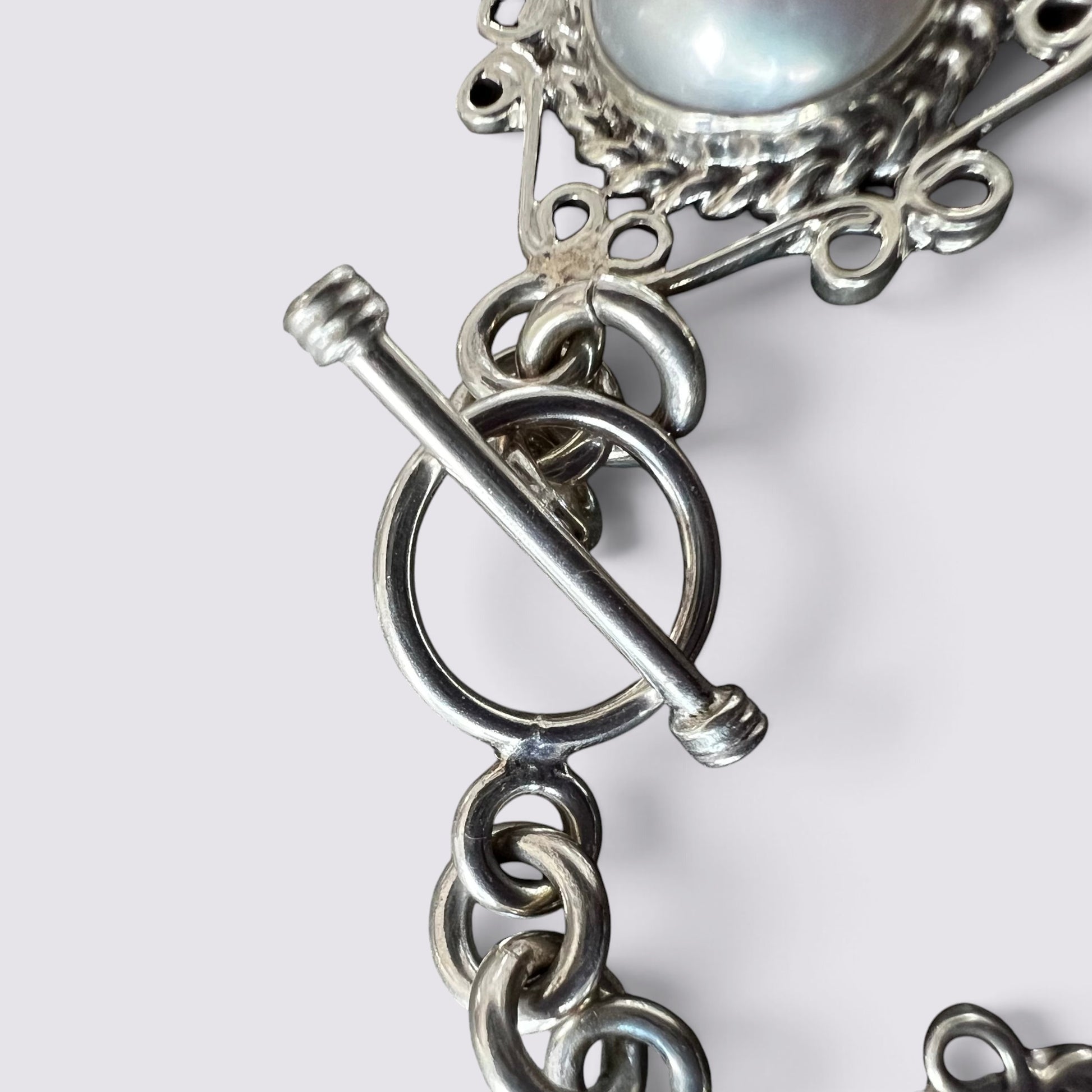 Close-up of a silver-toned chain bracelet featuring a toggle clasp and a round iridescent pearl at the top, shown against a light gray background.