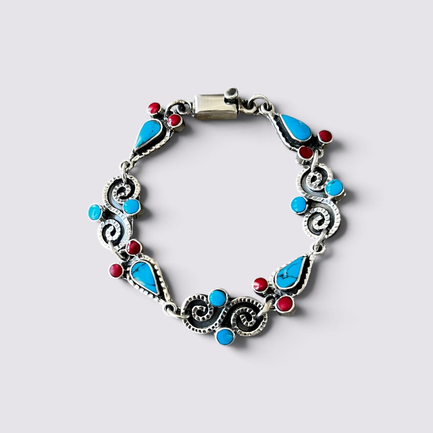 A silver-toned filigree bracelet featuring turquoise blue teardrop stones, small round blue stones, and red gemstone accents, displayed on a light gray background.