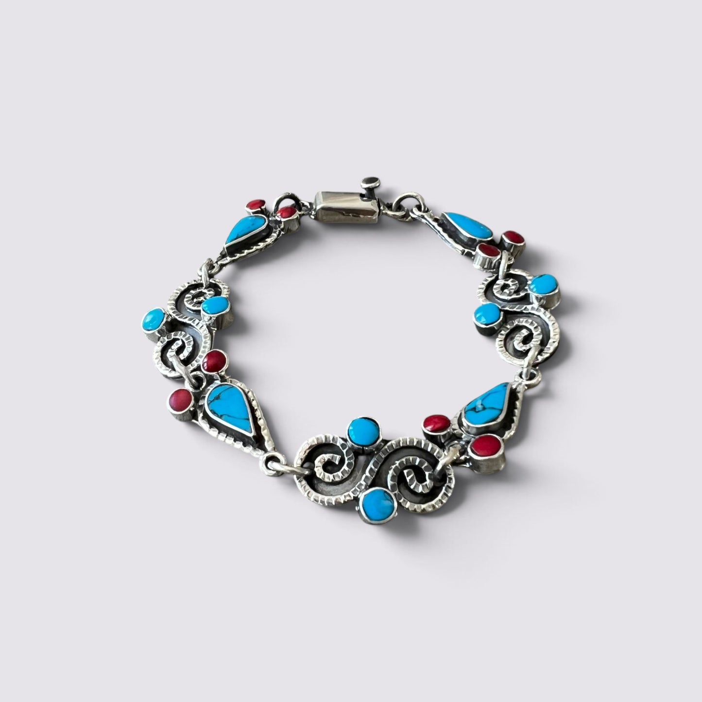A silver-toned bracelet featuring turquoise and red gemstone accents, displayed on a light gray background.