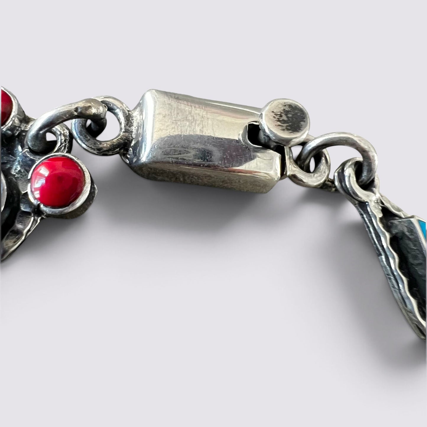 Close-up of a silver-toned bracelet clasp with red gemstone accents and a small turquoise detail, photographed on a light gray background.