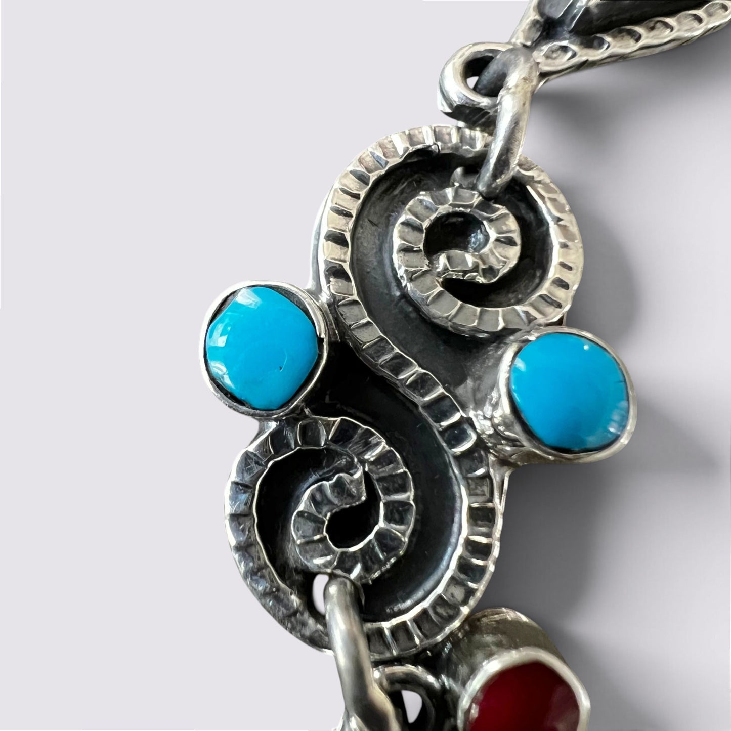 A close-up of a silver-toned spiral pendant necklace featuring two bright turquoise blue oval stones and a dangling red stone, set against a light gray background.
