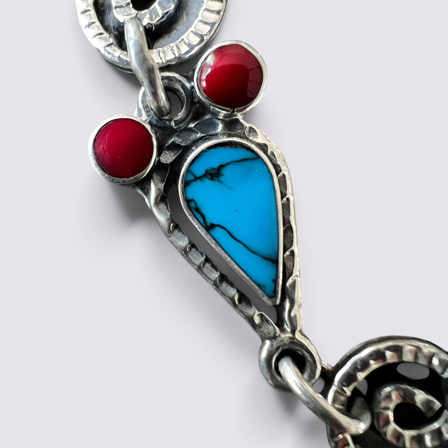 A close-up of a silver-toned jewelry charm featuring a turquoise blue teardrop-shaped stone inlay and two small round red stones, shown on a light gray background.