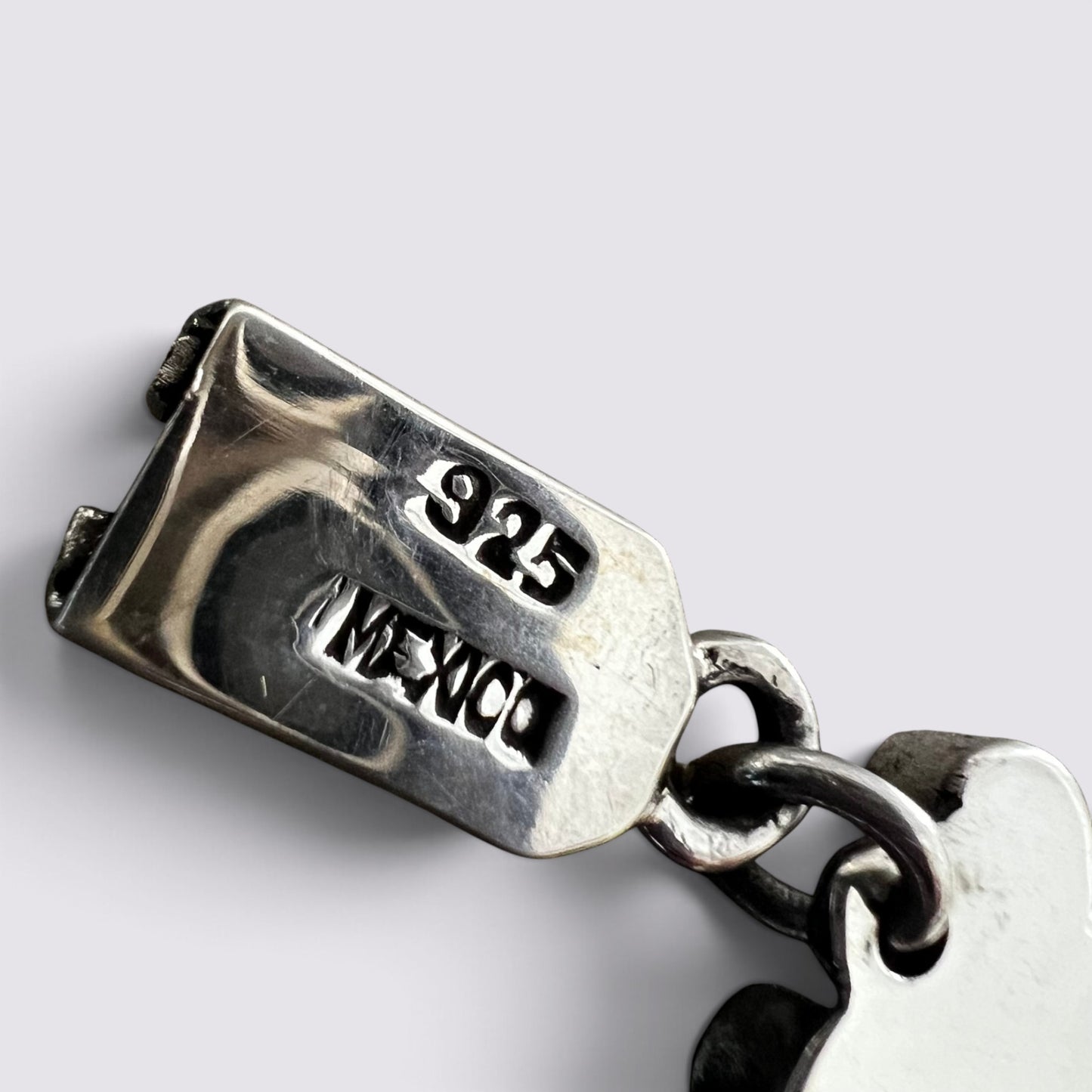 Close-up of a sterling silver charm link with '925' and 'MEXICO' engraved on the clasp, showing a polished metal surface and chain links against a light gray background.