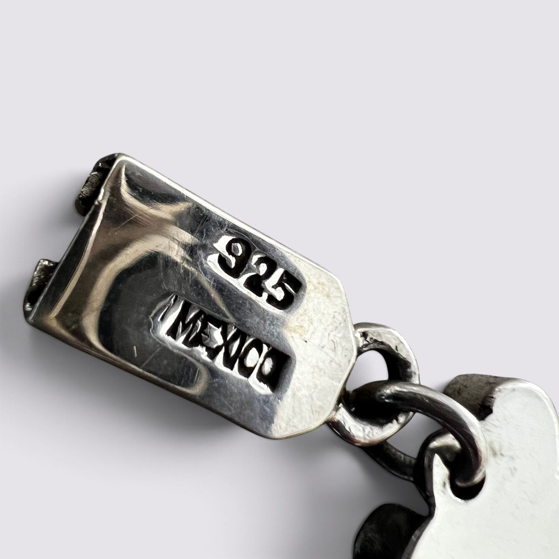 Close-up of a sterling silver charm link with '925' and 'MEXICO' engraved on the clasp, showing a polished metal surface and chain links against a light gray background.