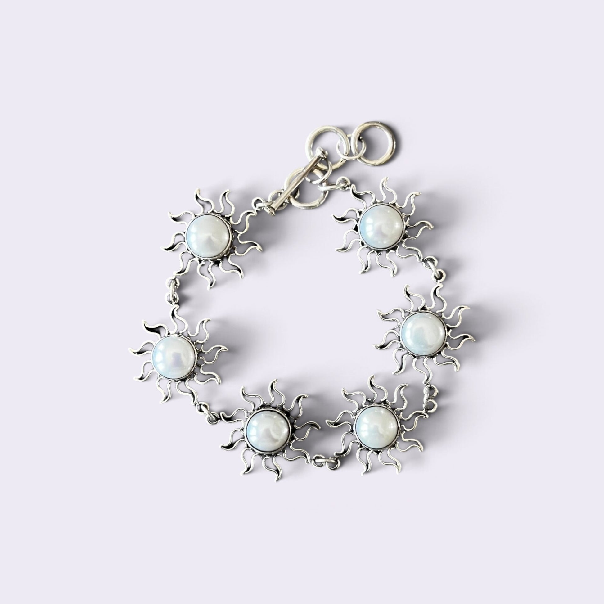 A silver-toned bracelet featuring six sunburst motifs, each with a round white faux-pearl center, arranged in a circle on a light lavender background.