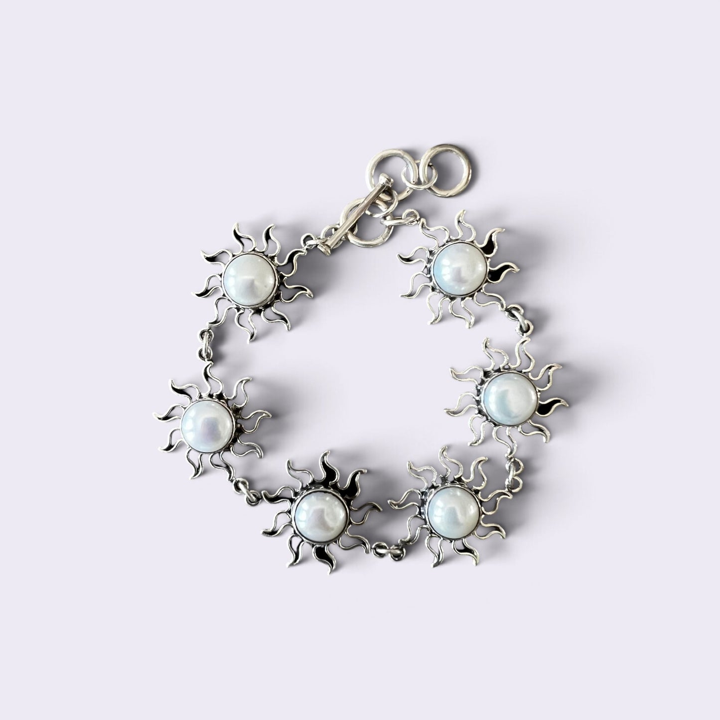 A silver-toned bracelet featuring six sunburst motifs, each centered with a round white faux pearl, arranged in a circle on a light lavender background.