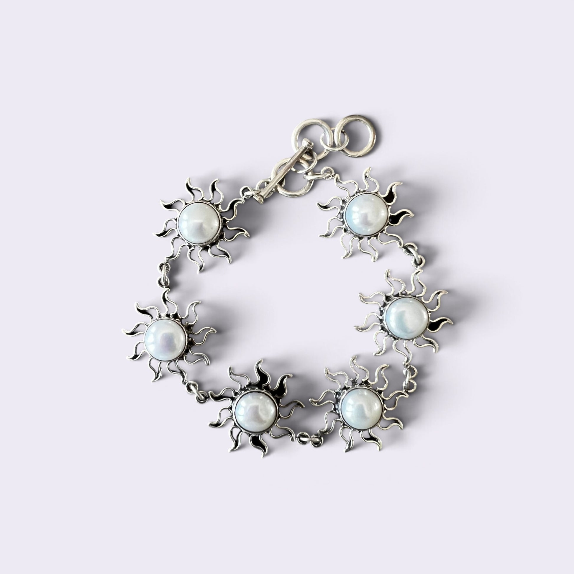 A silver-toned bracelet featuring six sunburst motifs, each centered with a round white faux pearl, arranged in a circle on a light lavender background.