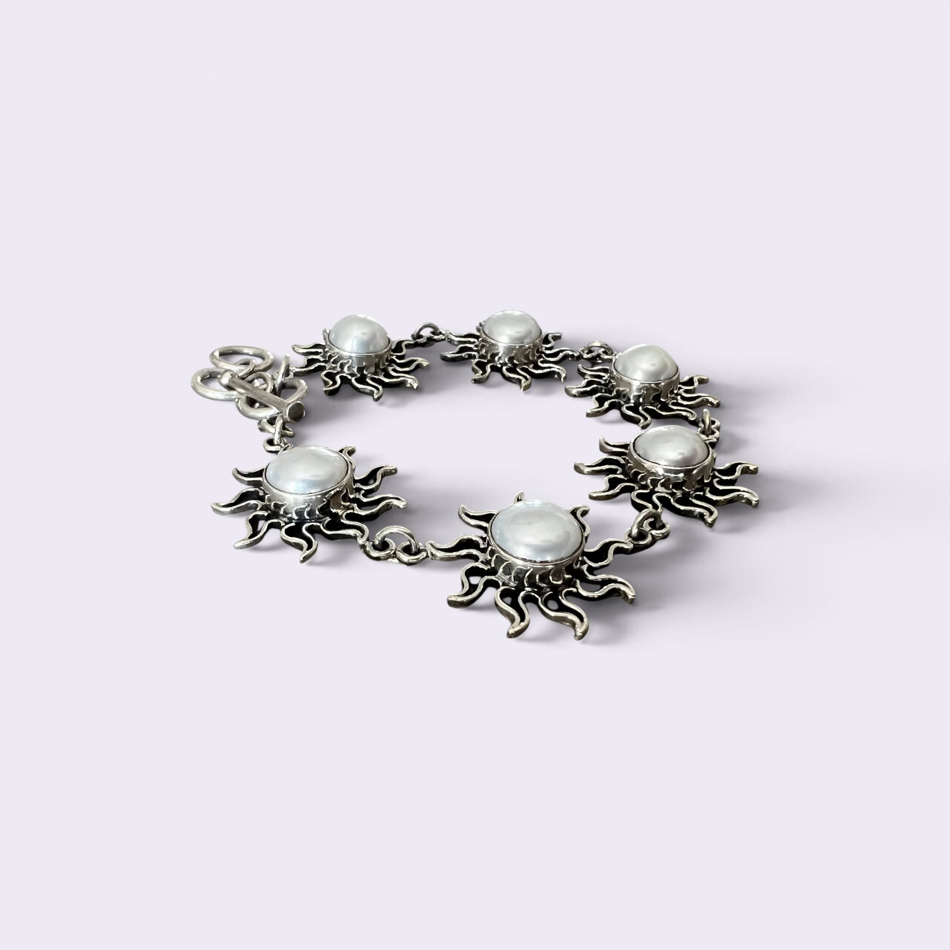 A silver-toned bracelet featuring five sunburst charms, each with a round white faux pearl in the center, arranged in a circle on a light lavender background.