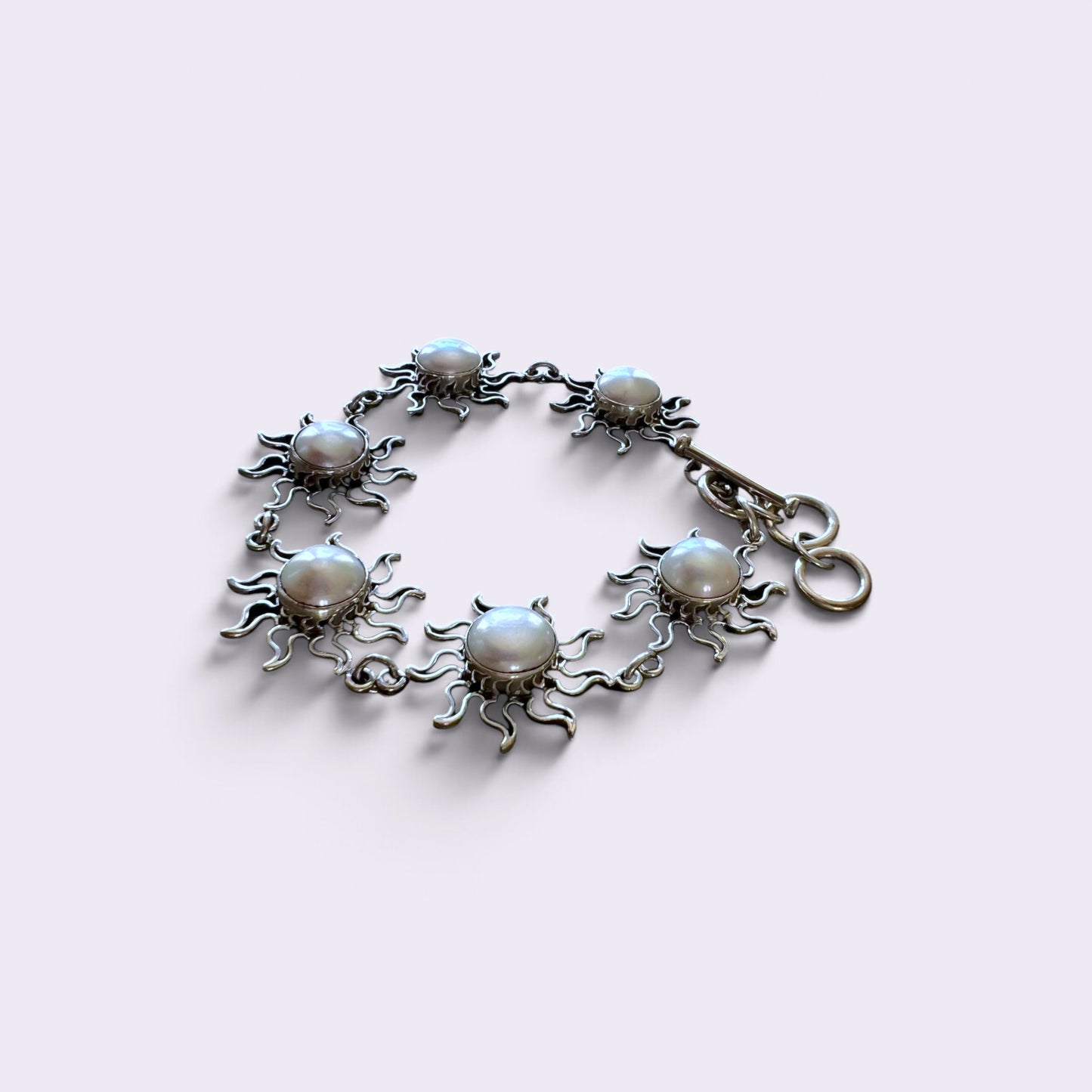 A silver-toned sunburst bracelet featuring seven round white faux pearls set in the center of each sun design, with a lobster clasp closure, shown on a light lavender background.