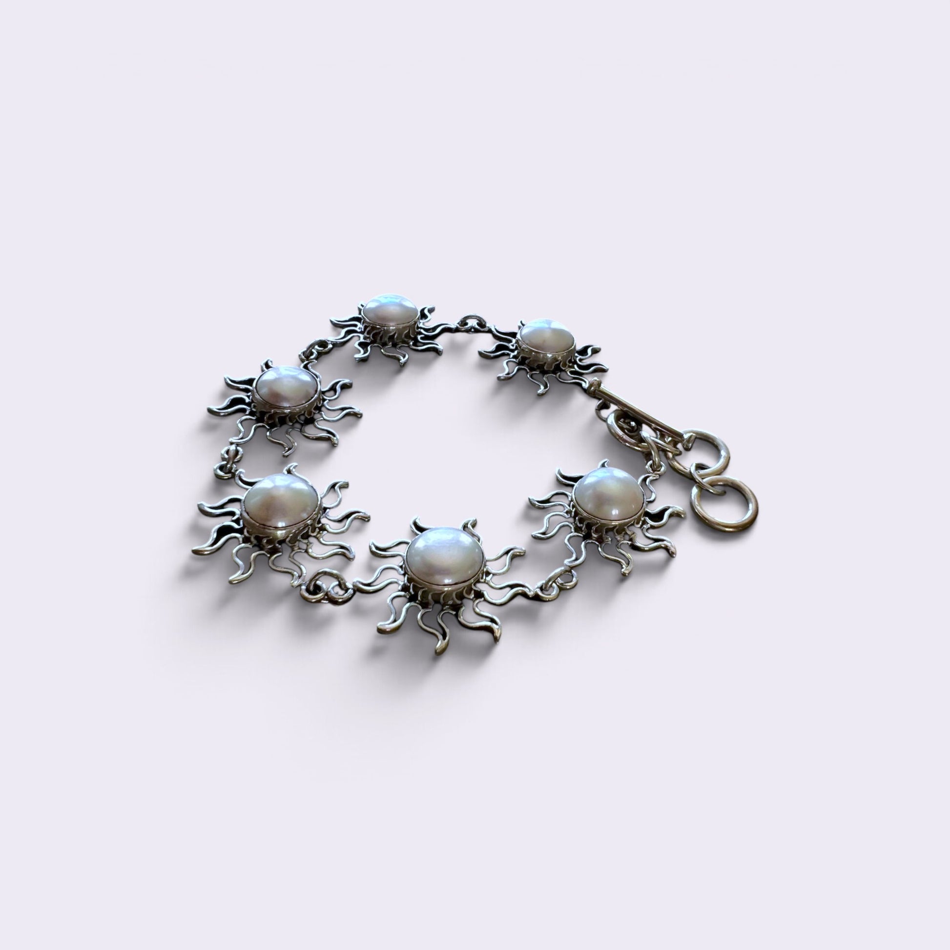 A silver-toned sunburst bracelet featuring seven round white faux pearls set in the center of each sun design, with a lobster clasp closure, shown on a light lavender background.