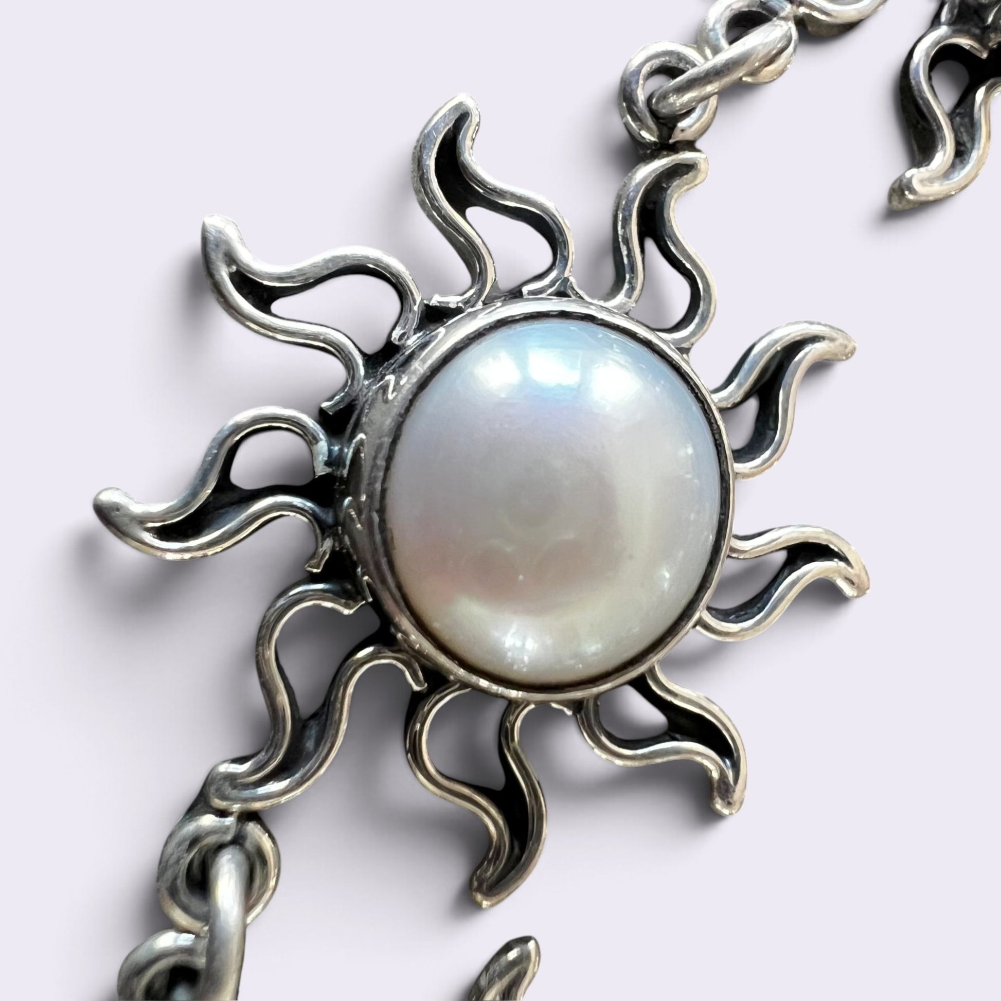 A close-up of a silver-toned sun pendant necklace featuring a large, round, iridescent white pearl in the center, set against a plain light background.