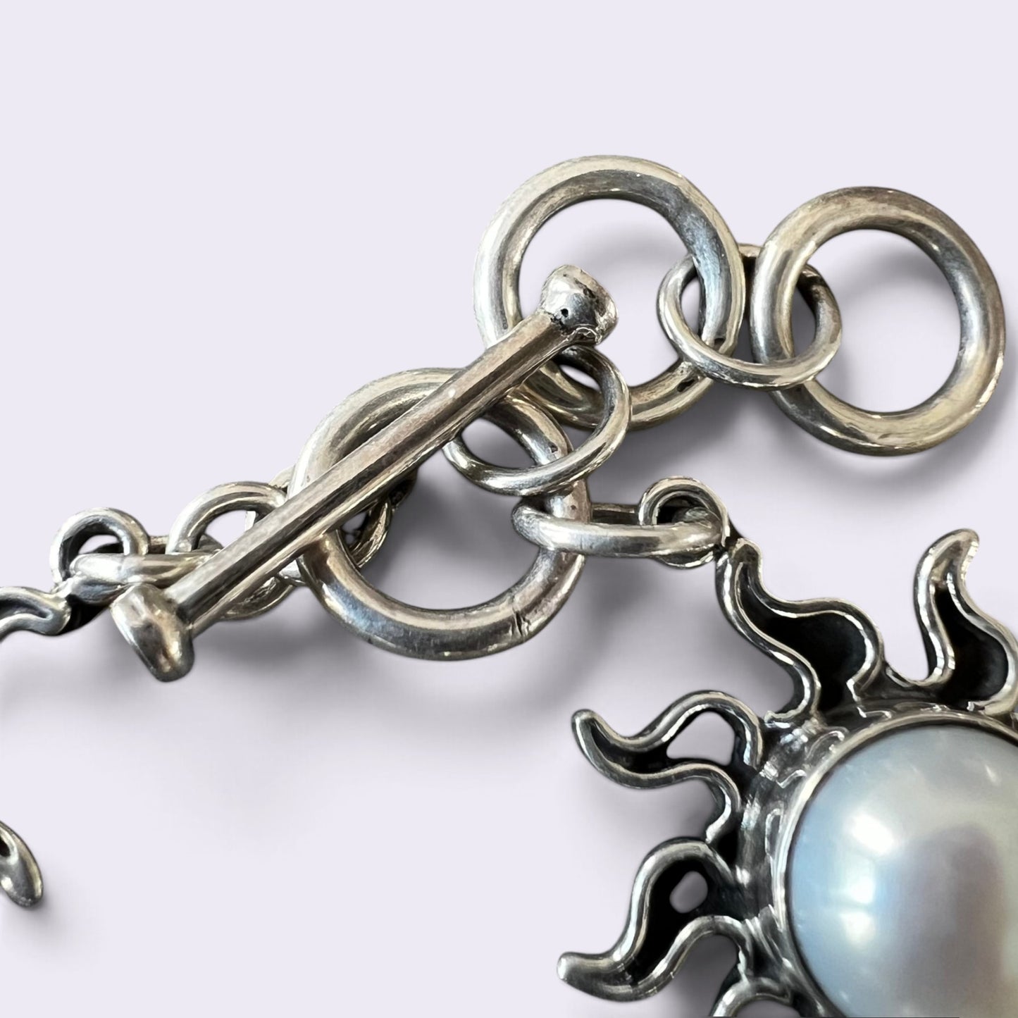 Close-up of a silver-toned jewelry piece with interlocking circular rings, a toggle clasp, and a sun-shaped charm holding a round white pearl, shown on a light gray background.
