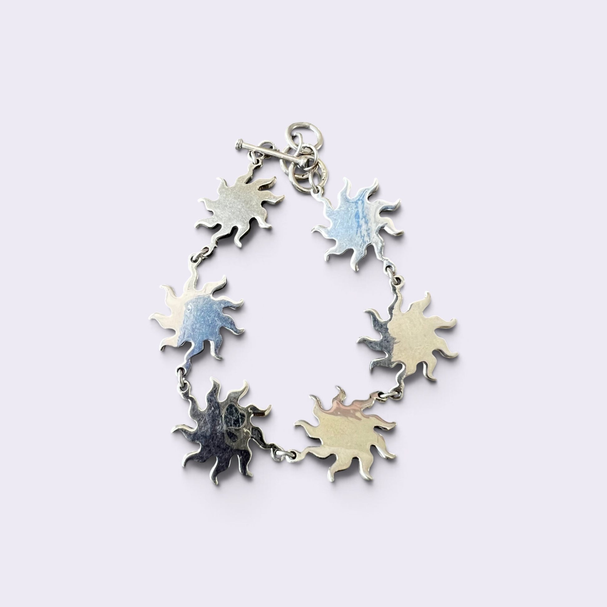 A silver-toned chain bracelet featuring seven irregular, sunburst-like charms with a hammered texture, displayed on a light lavender background.