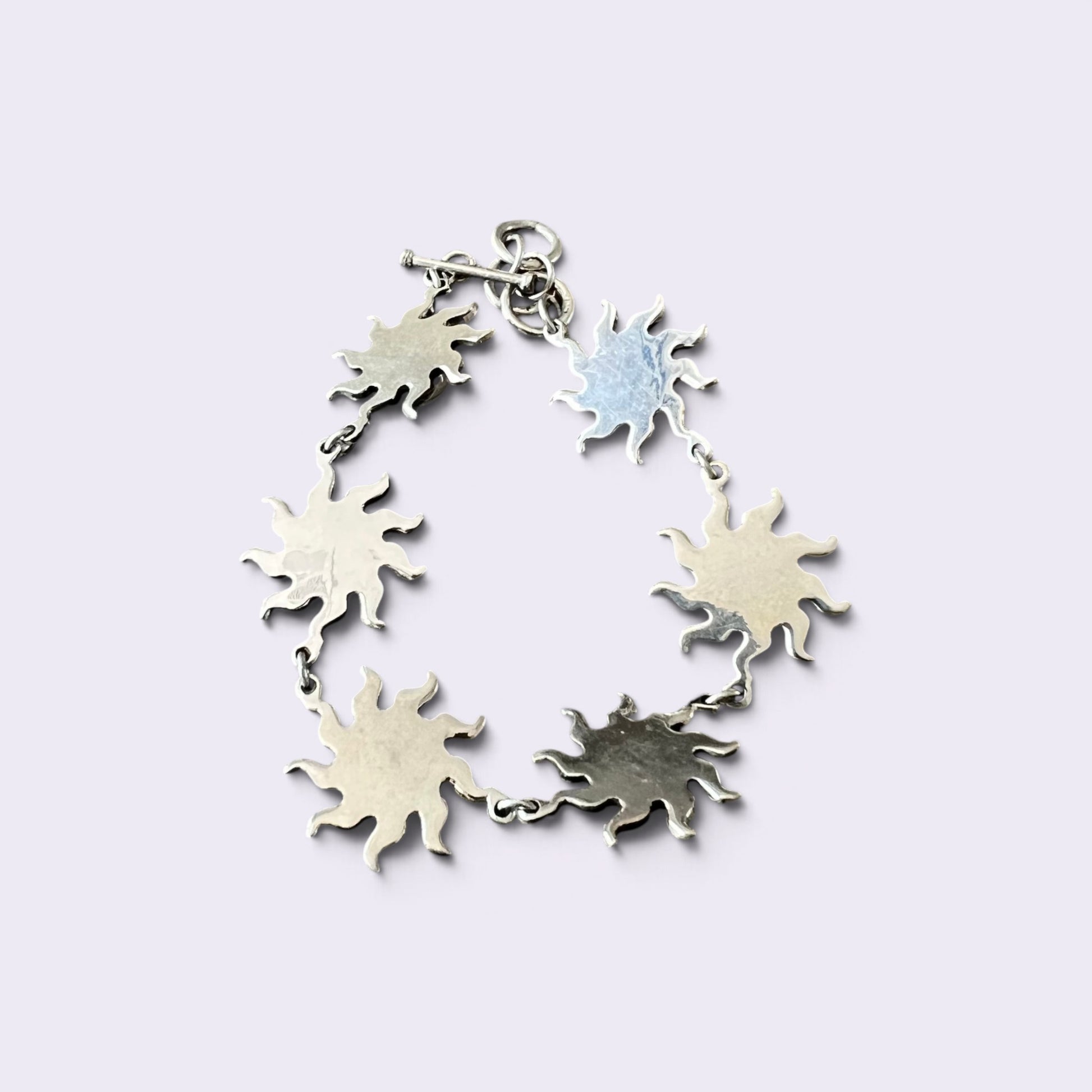A silver sunburst charm bracelet with five textured star-shaped charms connected by delicate silver chains, shown on a light lavender background.