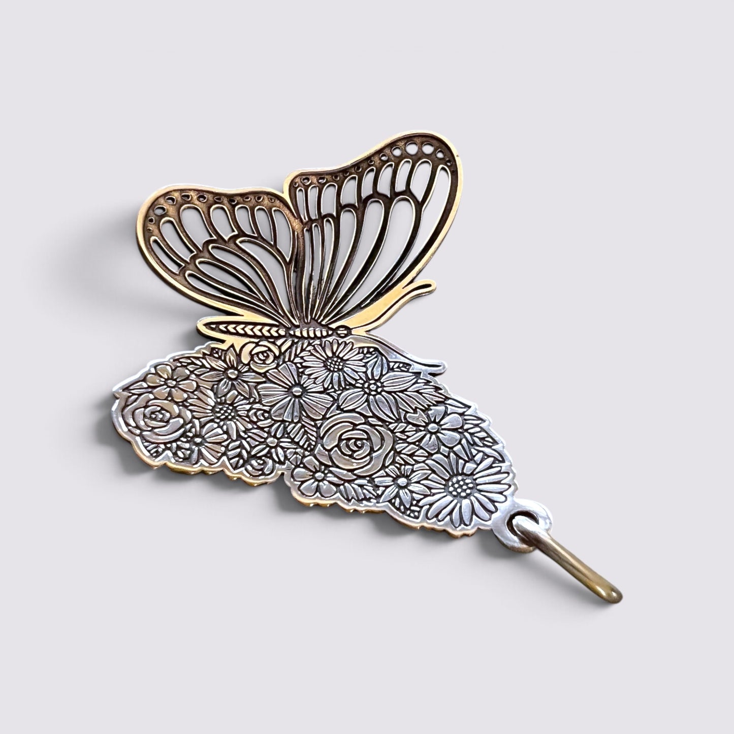 A butterfly-shaped brooch with an intricate floral pattern on the lower half and openwork wings on the upper half, finished in gold and silver-tone metal, shown on a light gray background.