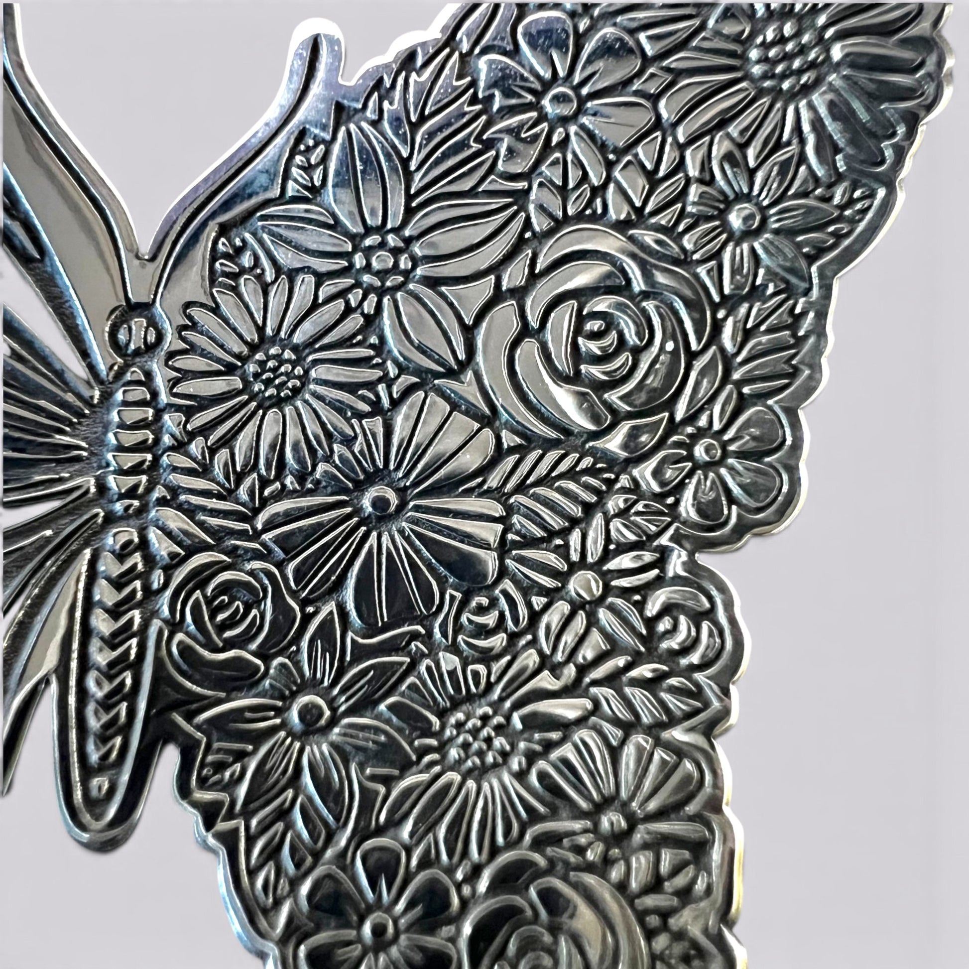 A close-up of a metallic butterfly-shaped decorative piece with an embossed floral pattern, including roses, daisies, and leaves, shown against a plain light gray background.