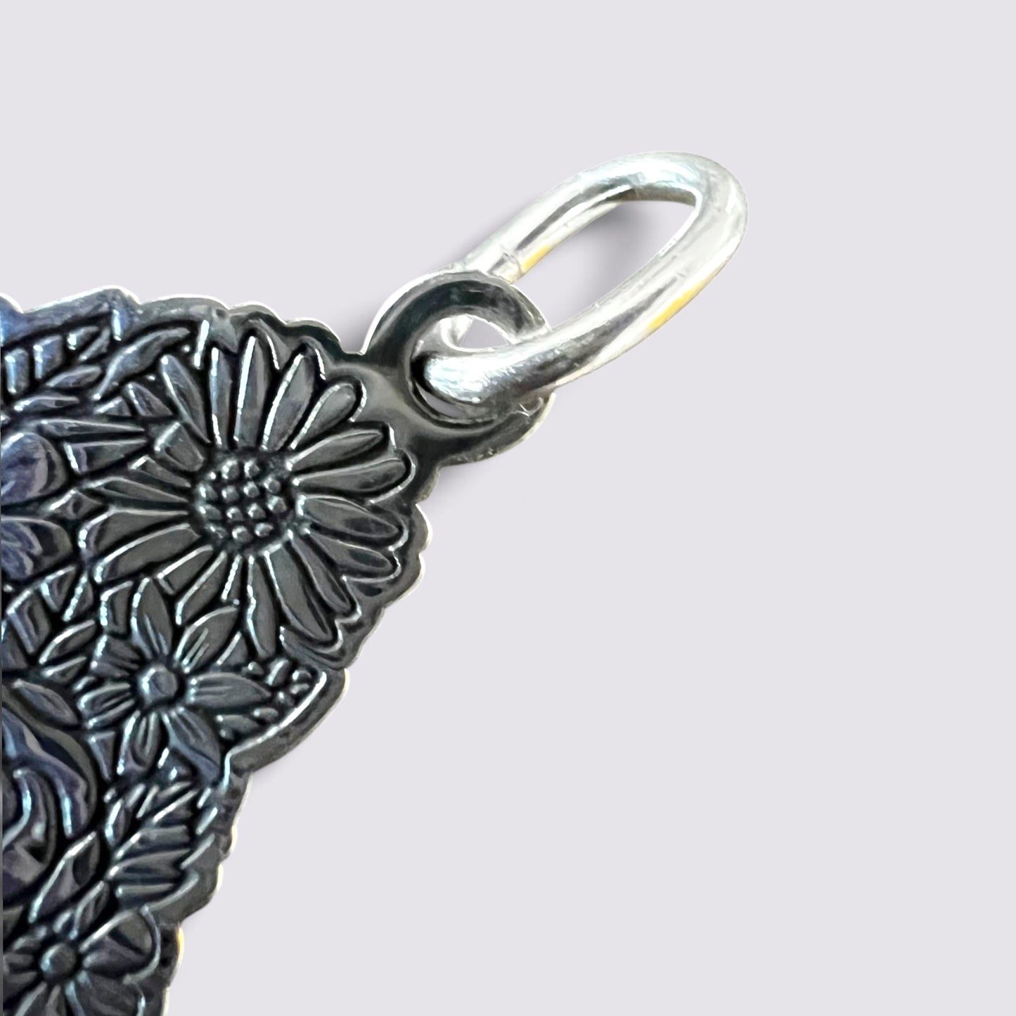 Close-up of a silver-toned metal charm with an embossed floral pattern and a loop at the top, shown against a light gray background.