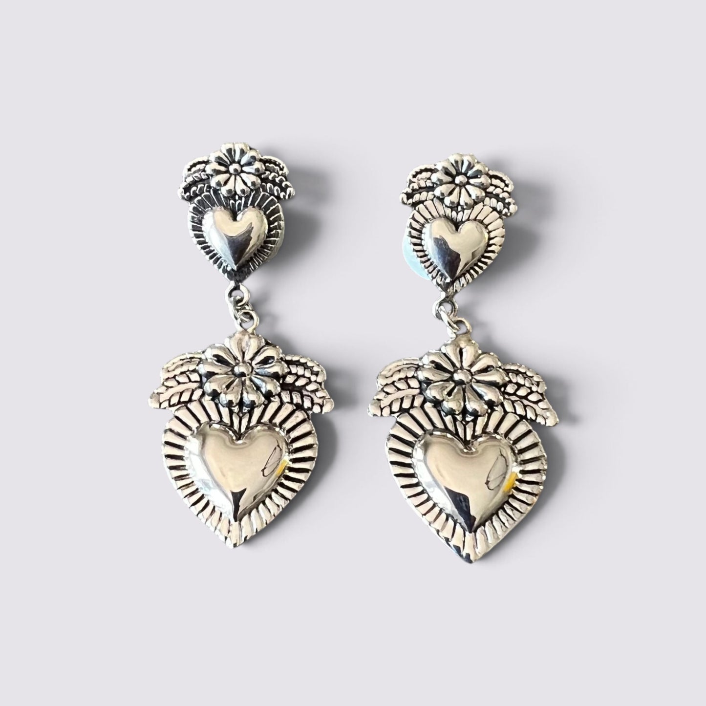 A pair of silver-toned dangle earrings with heart-shaped charms, floral details, and sunburst patterns, displayed on a light gray background.