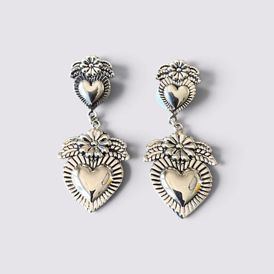 A pair of silver-toned dangle earrings with heart-shaped charms, floral details, and sunburst patterns, displayed on a light gray background.